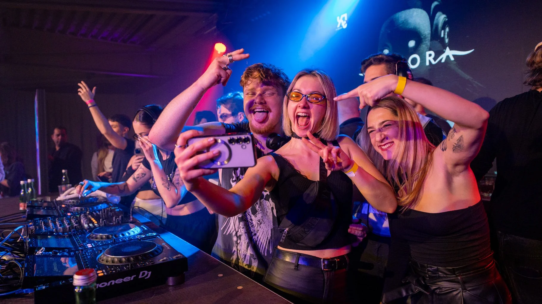 Menschen feiern bei einer Party in einem Club, sie machen Selfies und tanzen vor einem DJ-Pult, das im Hintergrund rot und blau beleuchtet ist.