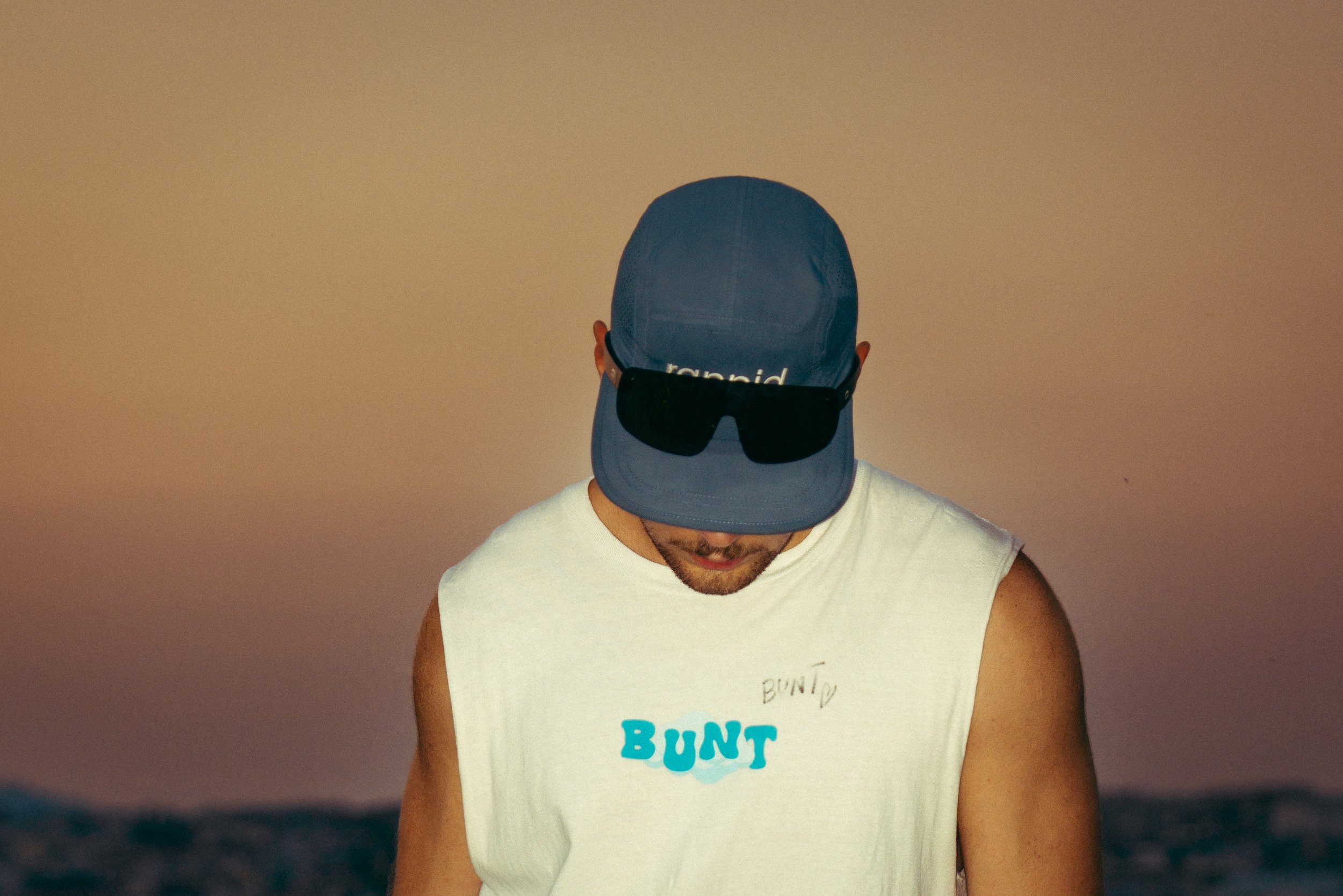 Mann trägt blaue Cap mit Sonnenbrille oben drauf, weißes Tanktop mit buntem Aufdruck, in einem Raum mit neutralem Hintergrund.