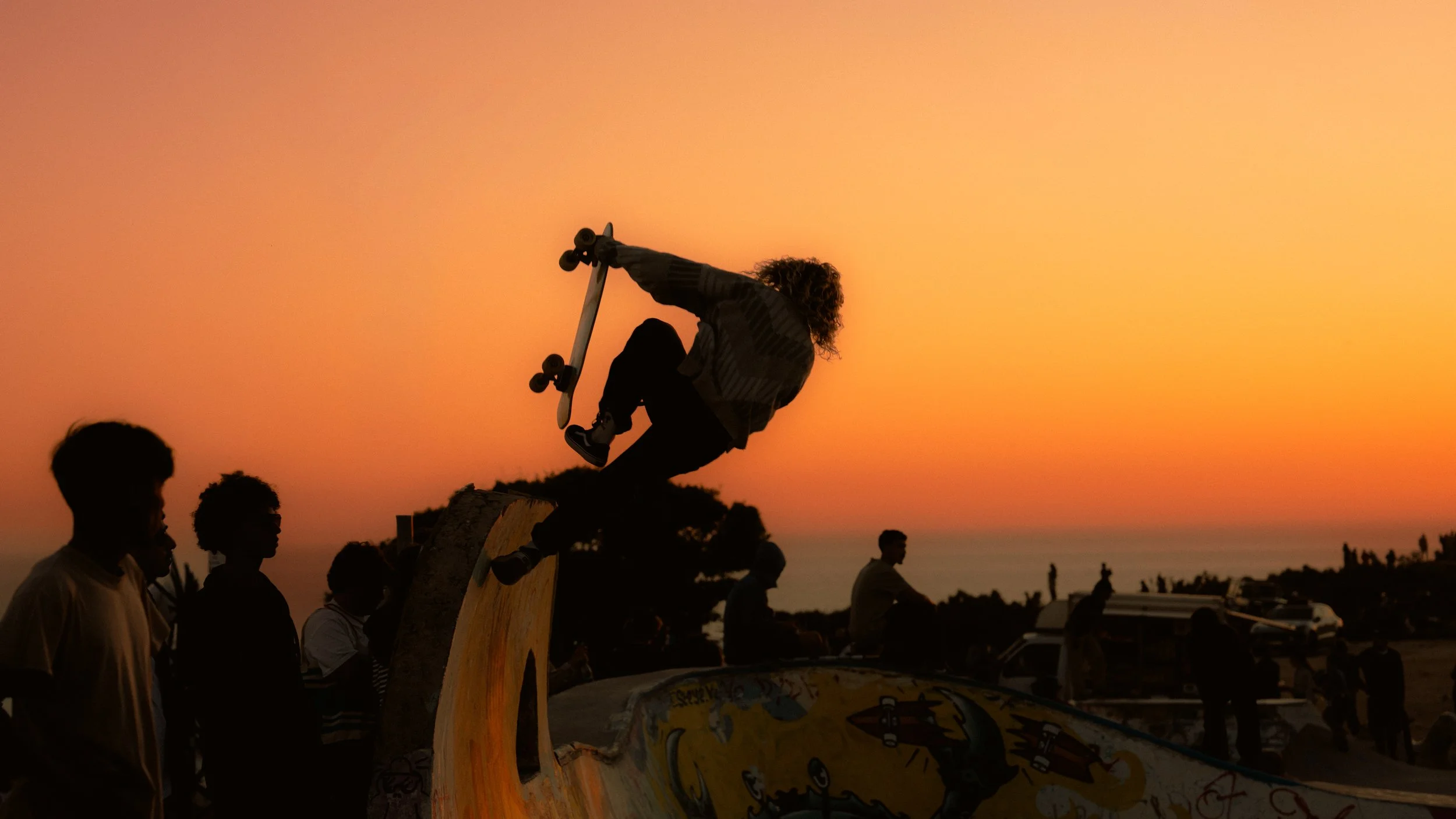 Eine Person springt bei Sonnenuntergang auf einem Skateboard auf einem Skatepark, während andere Menschen im Hintergrund zuschauen.