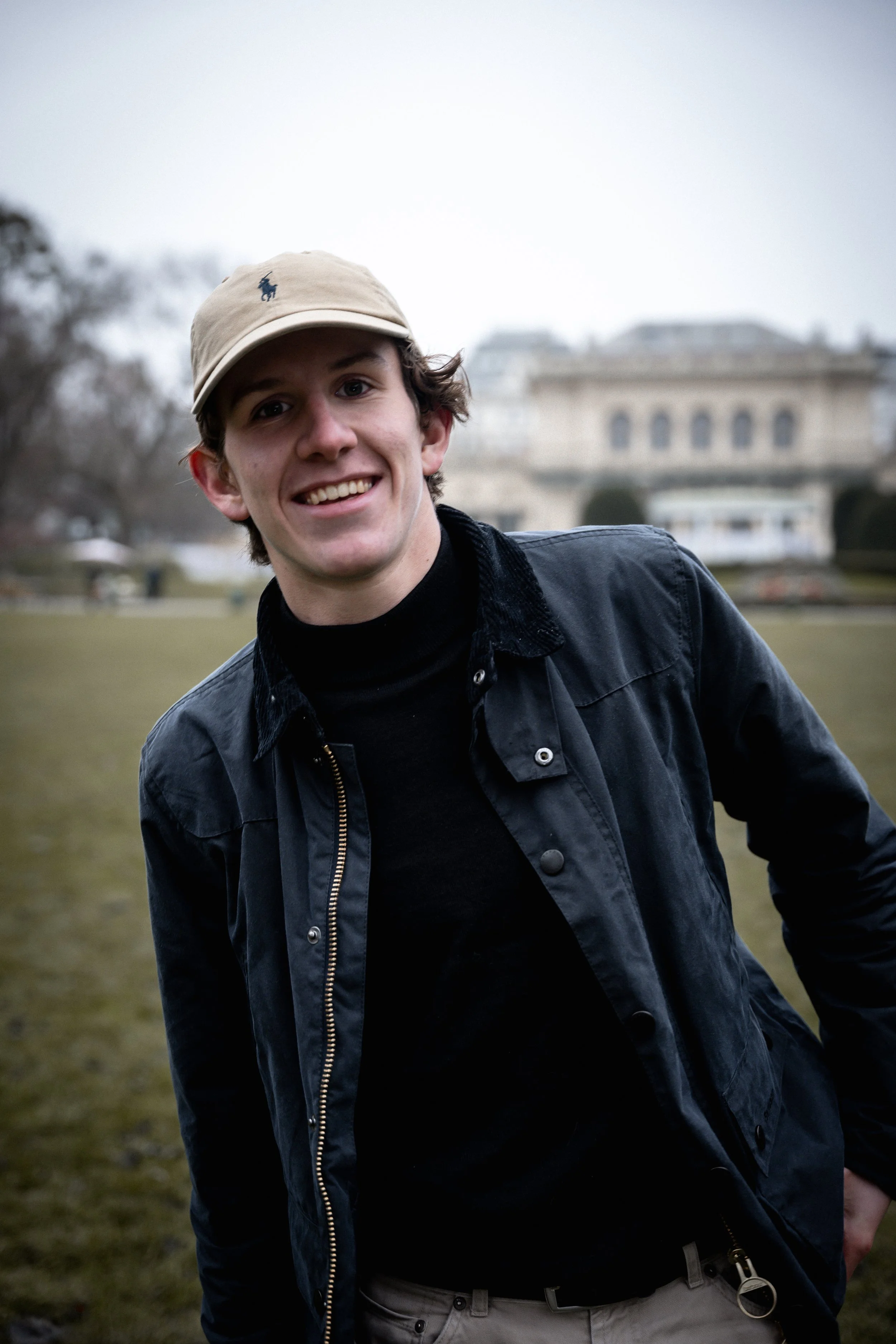 Junge Mann trägt beige Cap mit Pferd-Logo, lächelt, trägt schwarze Jacke und graue Hose, im Hintergrund ein Gebäude und Bäume bei bewölktem Himmel.