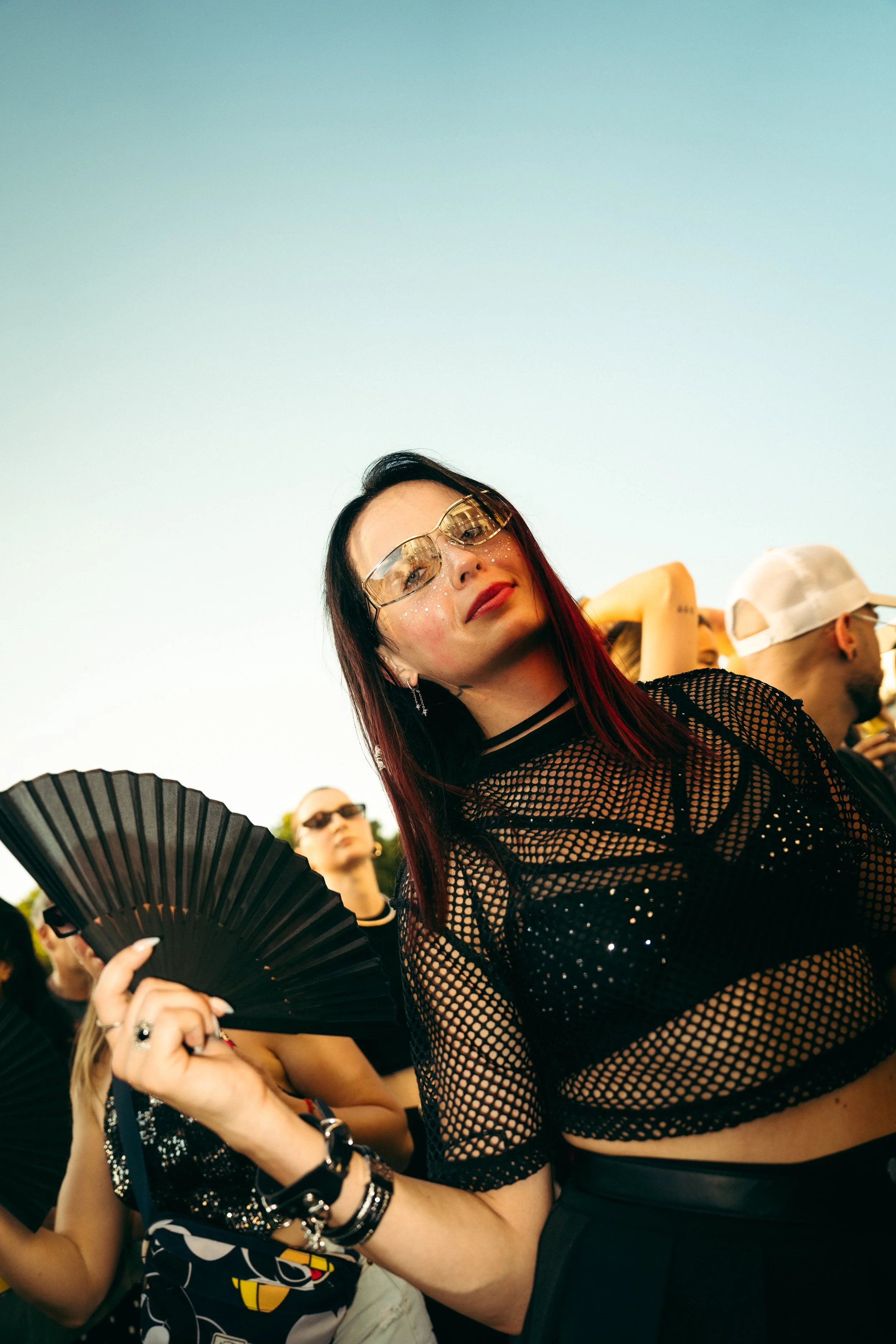 Junge Frau mit dunklen Haaren, Sonnenbrille und Netzshirt posiert bei einem Festival, umgeben von anderen Menschen, einige tragen Sonnenbrillen und typische Festivalkleidung, im Hintergrund ist der Himmel zu sehen.