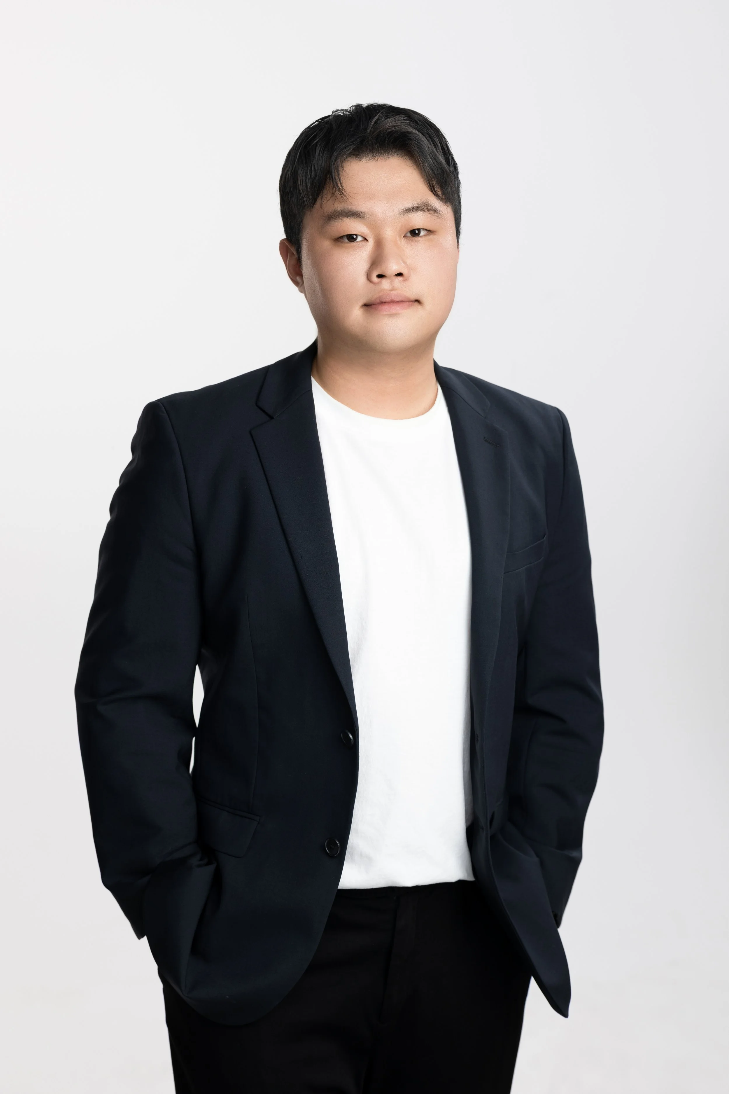 Portrait of a young man wearing a black blazer over a white t-shirt, standing against a plain light-colored background.