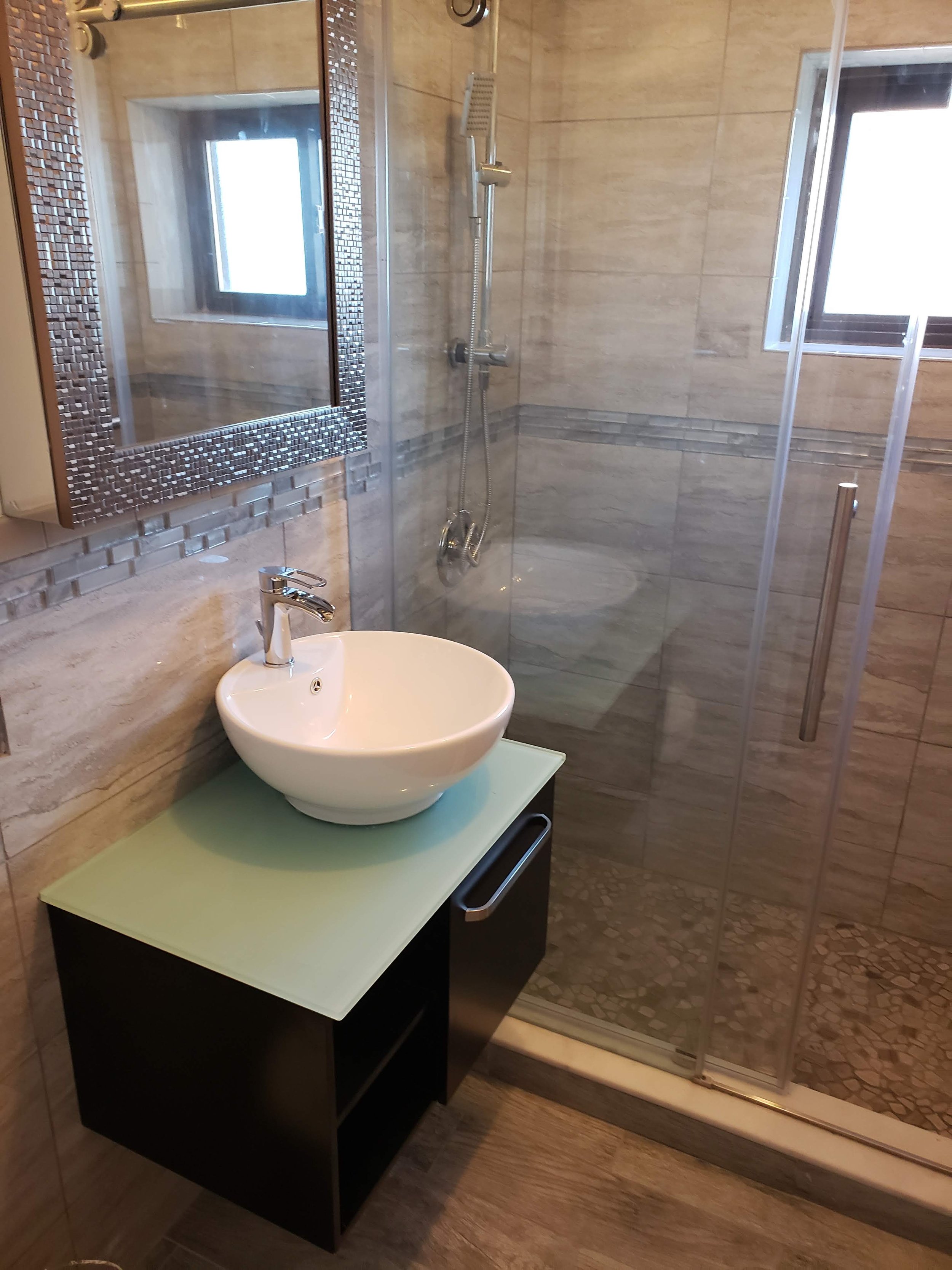 A modern bathroom with a round vessel sink on a black vanity with a glass top, a large rectangular mirror with a mosaic tile border, a window, and a walk-in shower with glass door and beige wall tiles.