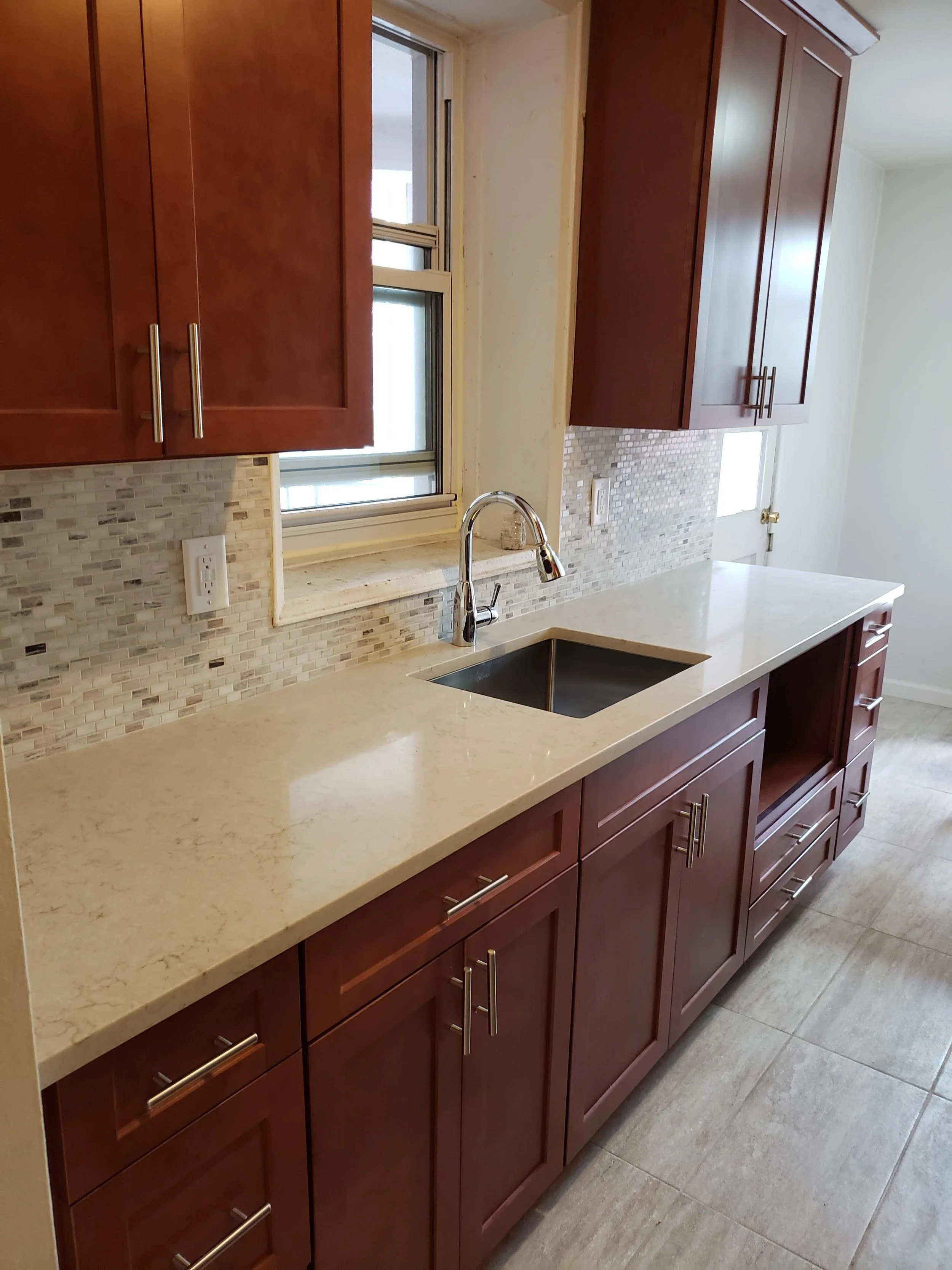 Kitchen with wooden cabinets, marble countertop, mosaic tile backsplash, window above the sink, and a door in the background.