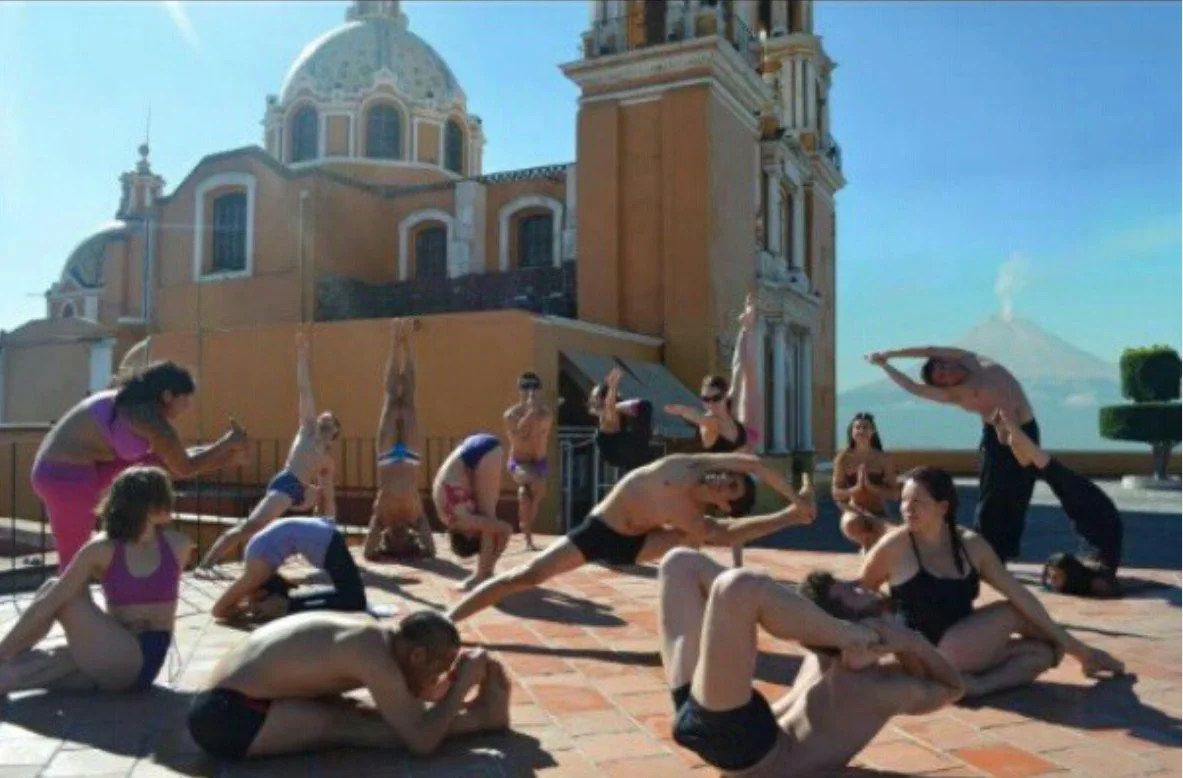 Group of people practicing yoga on a rooftop with historic buildings and volcano in the background during daytime.