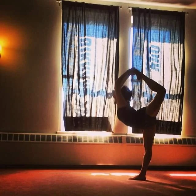 A person performing a yoga pose in a dimly lit room with curtains and a heater or radiator below the window.