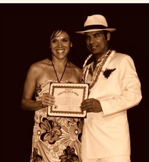 A woman in a strapless dress holding a certificate, standing next to a man in a light-colored suit and hat, in front of a dark background.