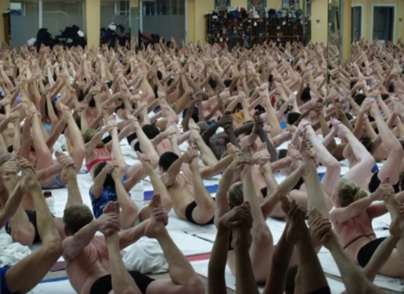 A large group of people practicing yoga indoors, sitting on mats with their arms raised and hands clasped together overhead.