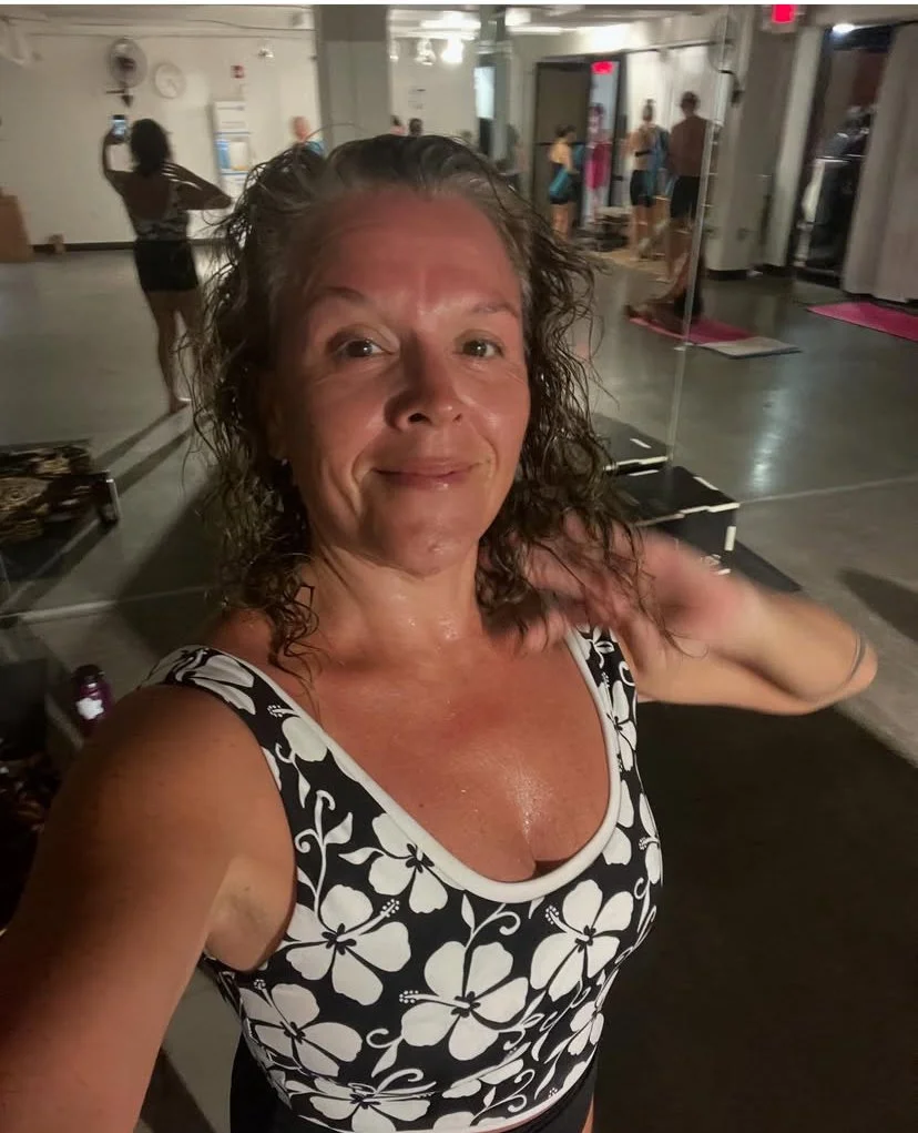 A woman takes a selfie in a fitness studio after a workout, with other people exercising and mirrors in the background.