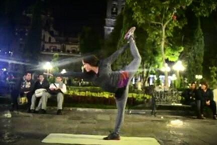 A person performing a yoga or dance pose outdoors at night, balancing on one leg with the other leg raised behind them and held with one hand, in a park or public space with benches and trees.