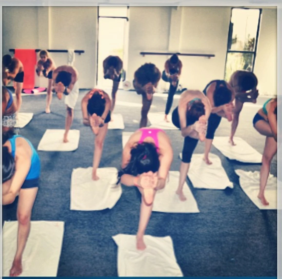 People participating in a yoga class, performing forward bends on yoga mats in a bright studio with large windows.