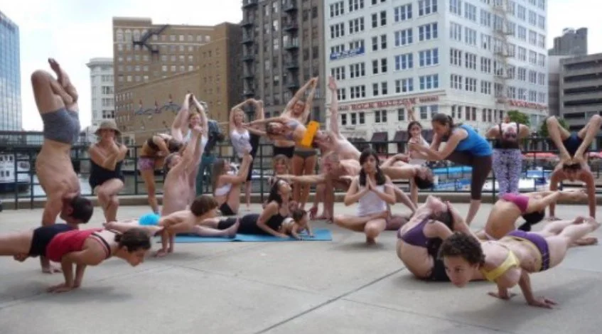 Group of people practicing yoga outdoors on a city rooftop, performing various poses on yoga mats with city buildings in the background.