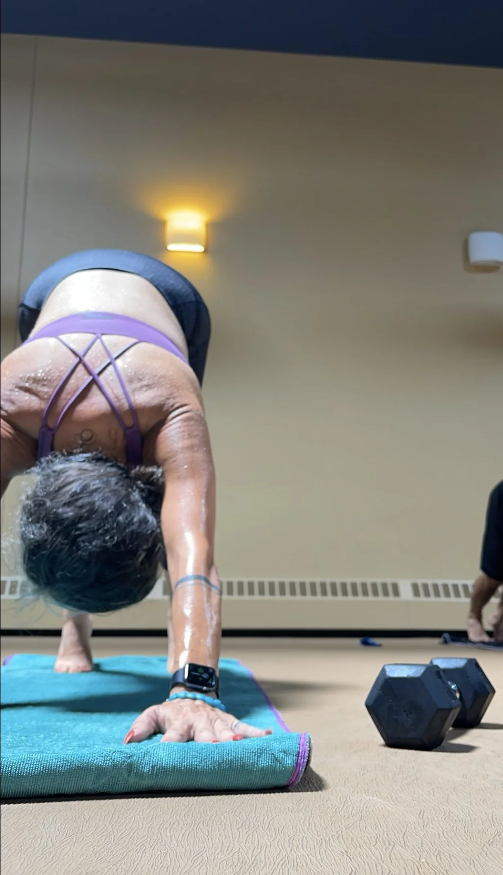 A woman practicing yoga in a room, seen from the front, performing a downward dog pose with her hands on a yoga mat, wearing a purple sports bra and shorts, with a fitness tracker on her wrist and dumbbells on the floor nearby.