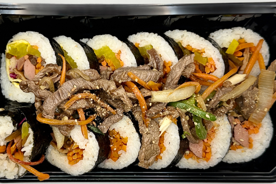 Korean kimbap rolls filled with rice, sausage, imitation crab, cucumber, carrot, pickled radish, fish cake, sesame oil, and sesame seeds, sliced and served fresh. Complemented with a side of marinated grilled beef.