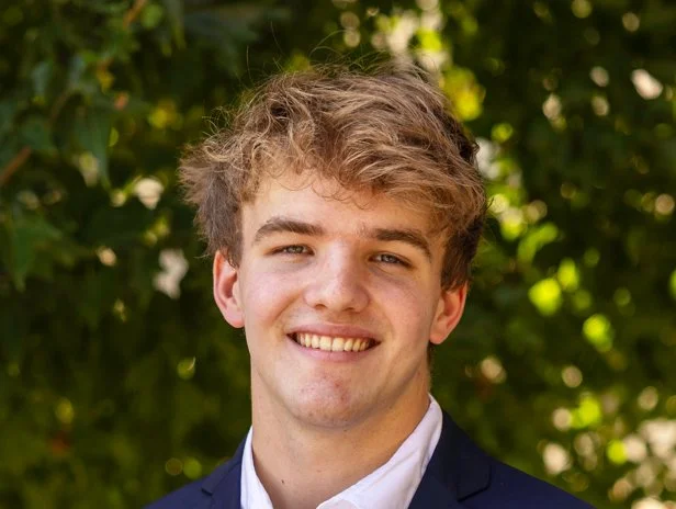 Young man in suit smiling outdoors with green trees in background.