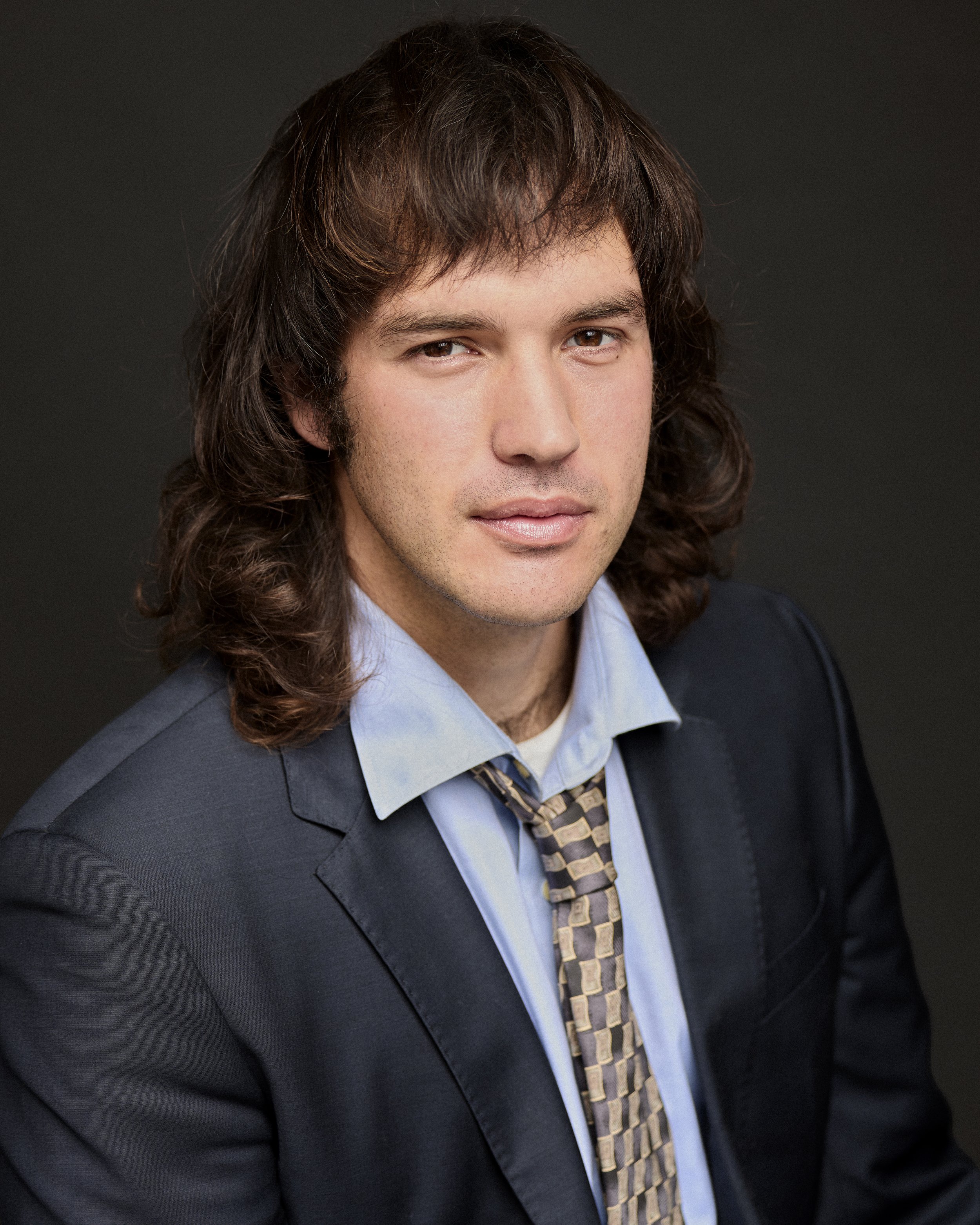 Photograph of a man wearing a business suit, dress shirt, and tie, with dark, long hair, against a black background.