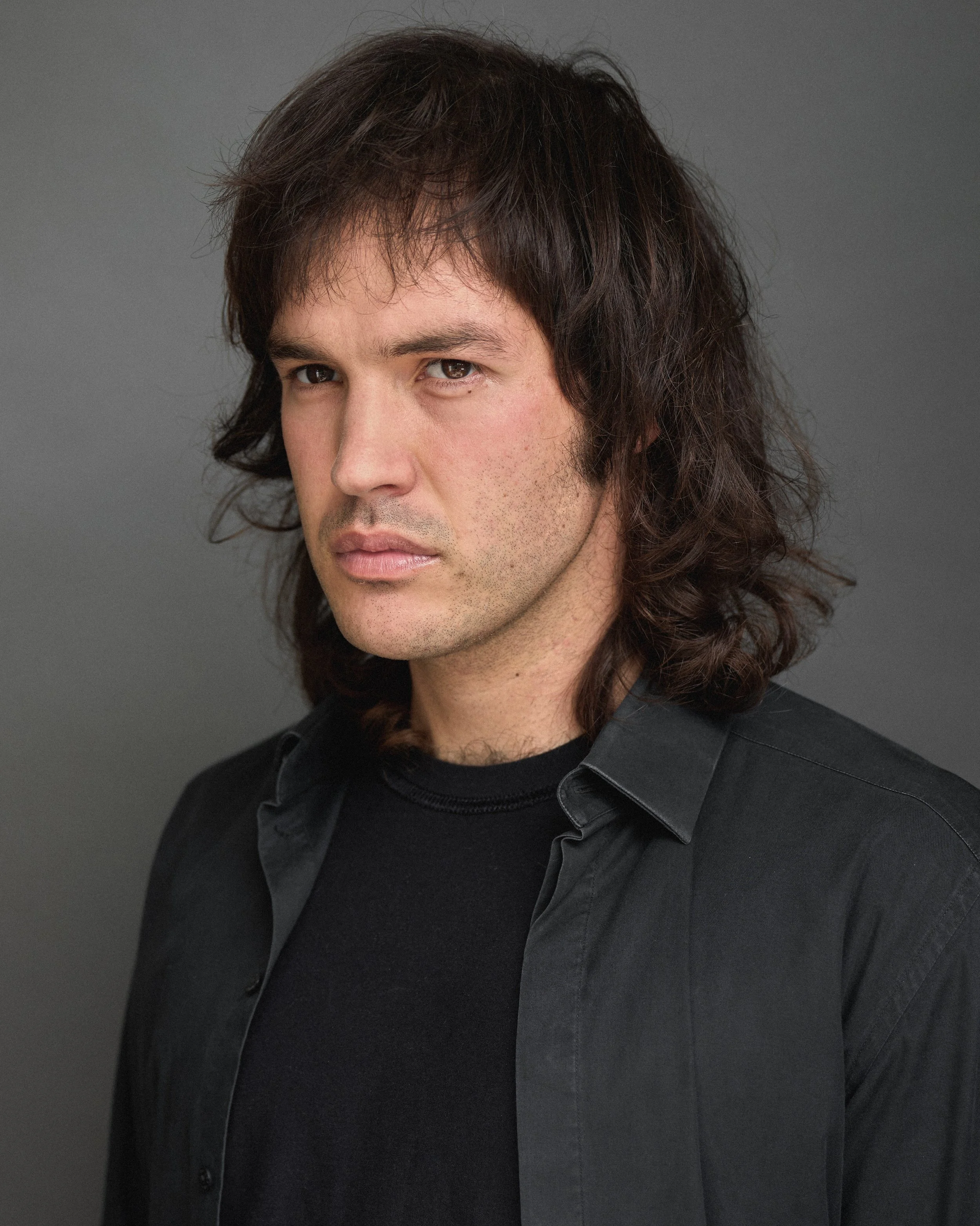 Close-up of a man with medium-length, wavy dark hair and fair skin, wearing a black shirt, against a gray background.