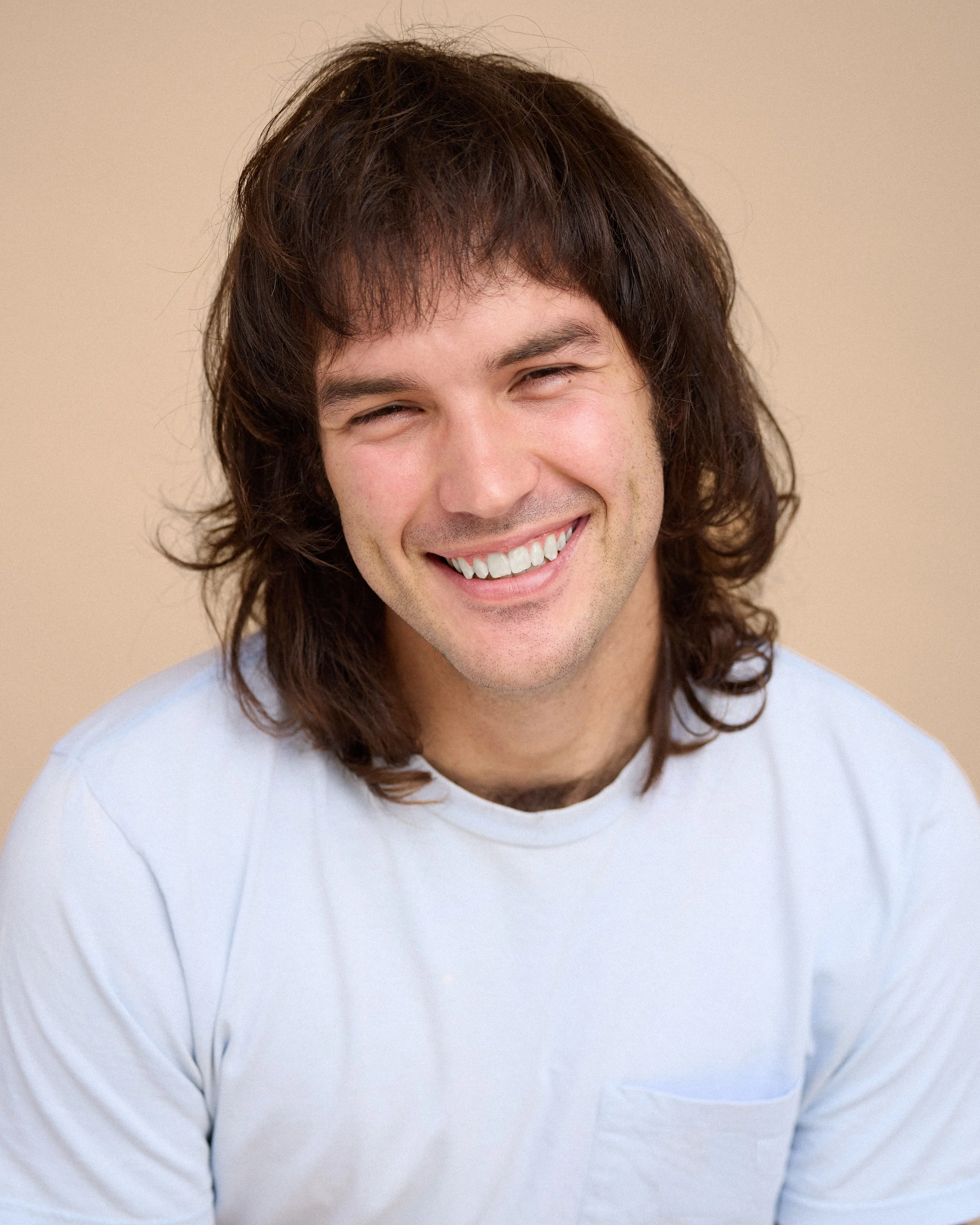 A young man with long brown hair, smiling broadly, wearing a white shirt, against a plain beige background.