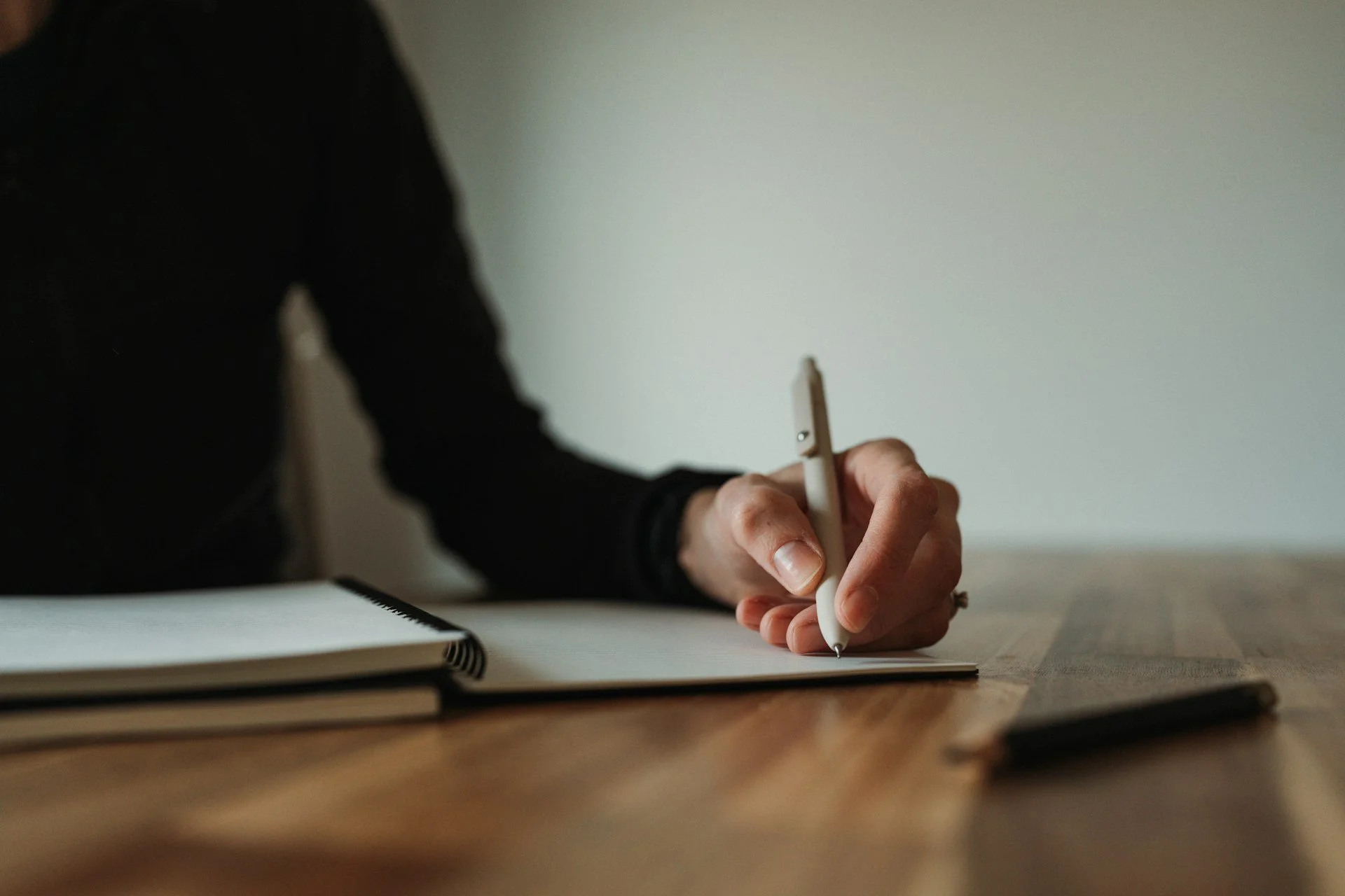 A person in a black long-sleeve shirt writing in a notebook with a pen on a wooden table, with a blank wall in the background.