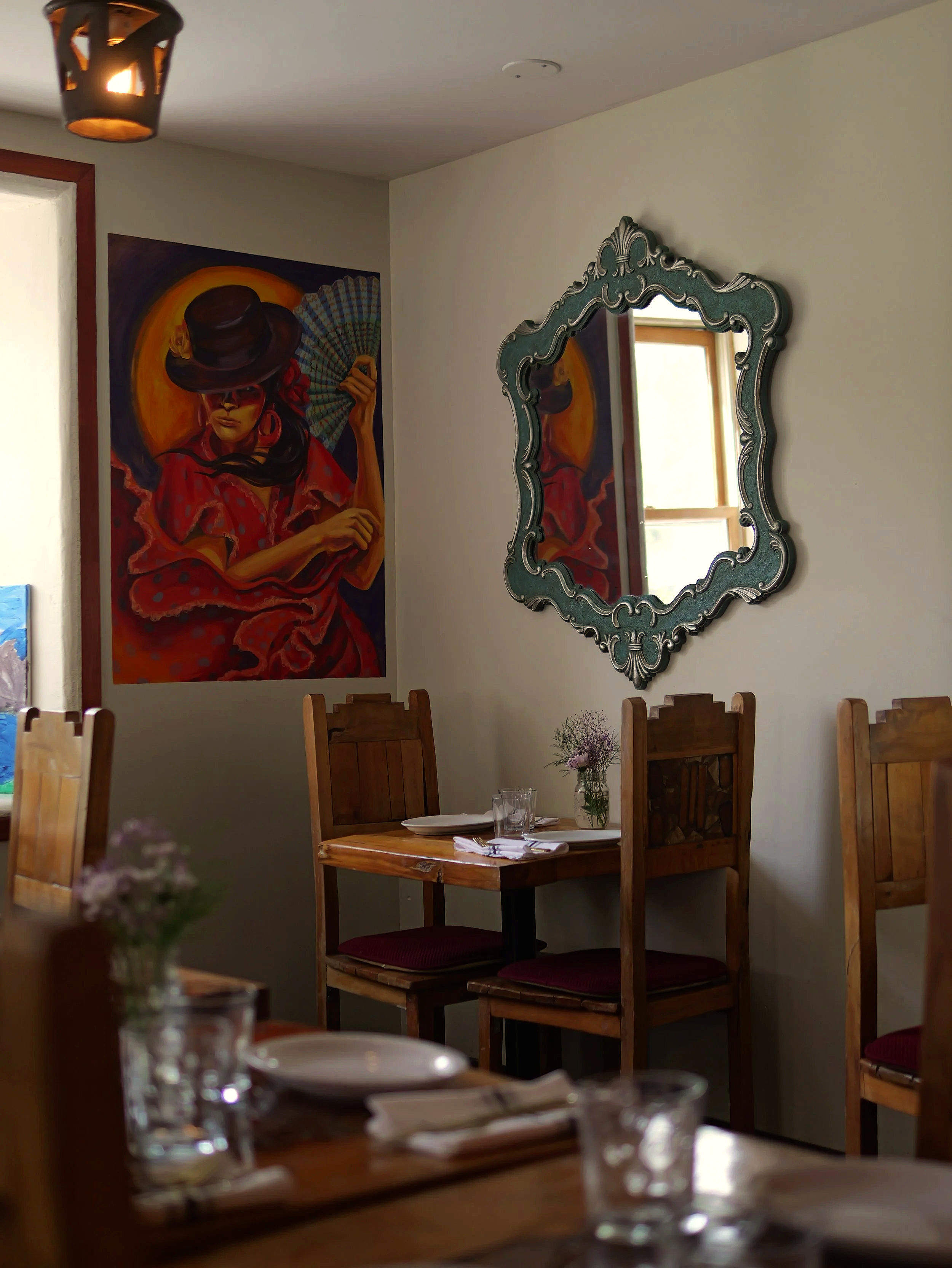 Intérieur d'un restaurant avec une peinture colorée sur le mur représentant une femme portant un chapeau et un éventail, un miroir orné, et des tables en bois avec assiettes, verres et fleurs.