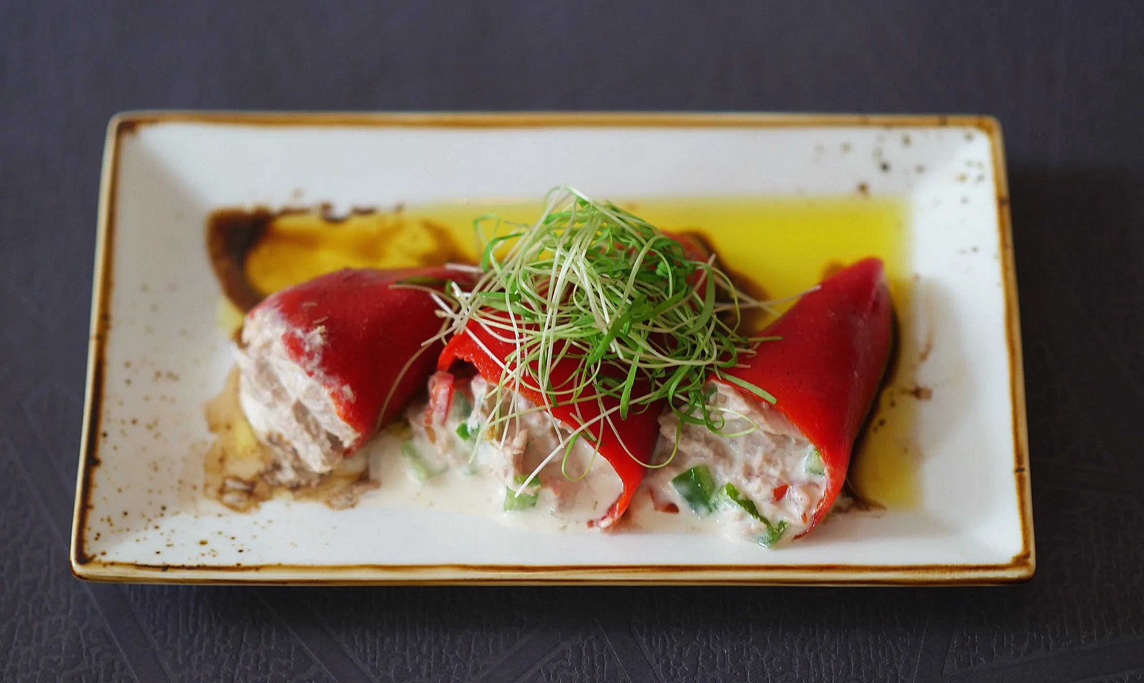 Un plat de poivrons avec de la garniture, de la crème, des oignons verts, et des germes, servi dans une assiette rectangulaire blanche avec de l'huile d'olive autour, Tapas