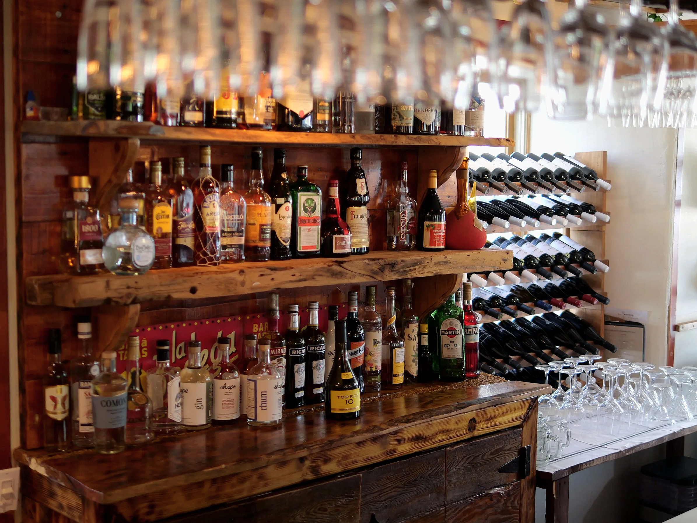 Une étagère en bois contenant diverses bouteilles d'alcool, y compris du gin, du tequila, du whisky, et d'autres spiritueux, ainsi que des rangées de bouteilles de vin et des verres à vin sur un comptoir en bois.