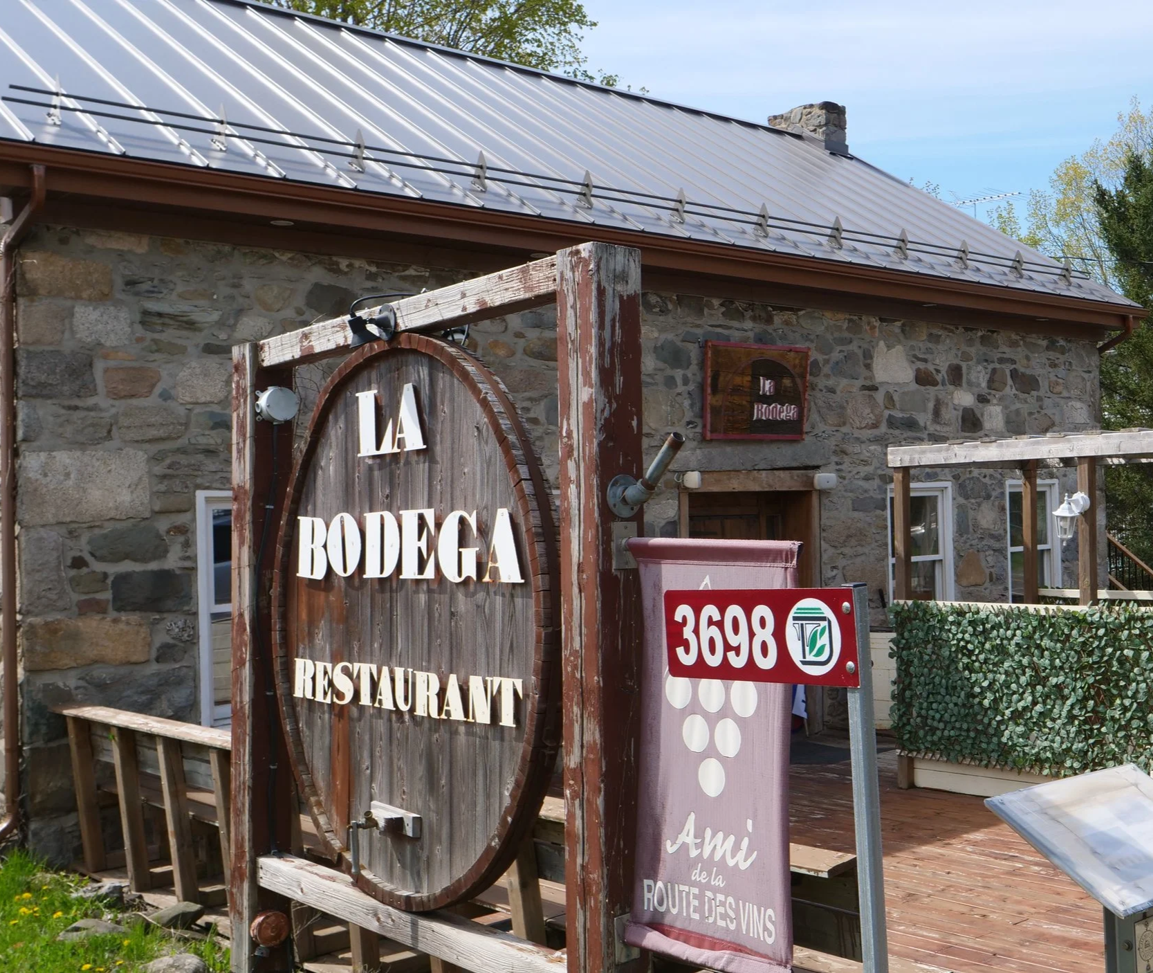 Un bâtiment en pierre avec une enseigne en bois annonçant une bodega et un restaurant. Il y a des panneaux et des décorations extérieures, y compris un panneau avec le numéro 3698 et une affiche rose avec le texte 'Ami de la route des vins'.