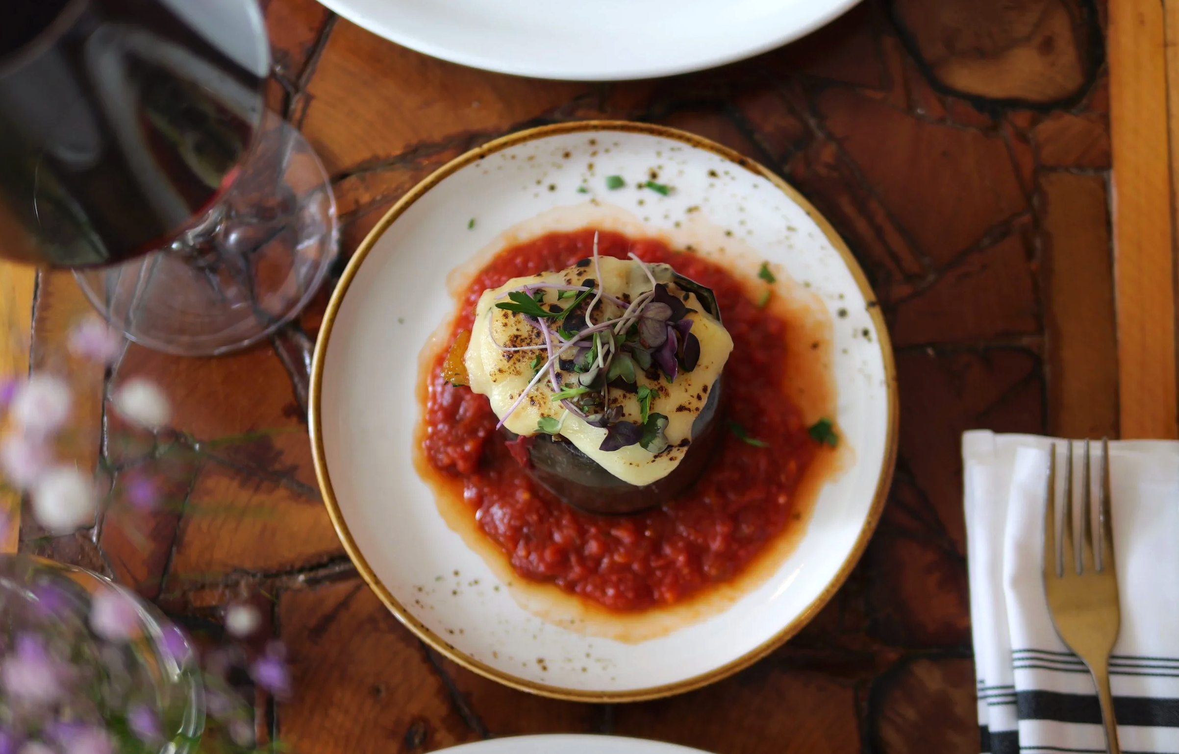 Assiette avec un aubergine farcie, garnie de microgreens, sur une sauce tomate, accompagnée d'un verre de vin rouge.