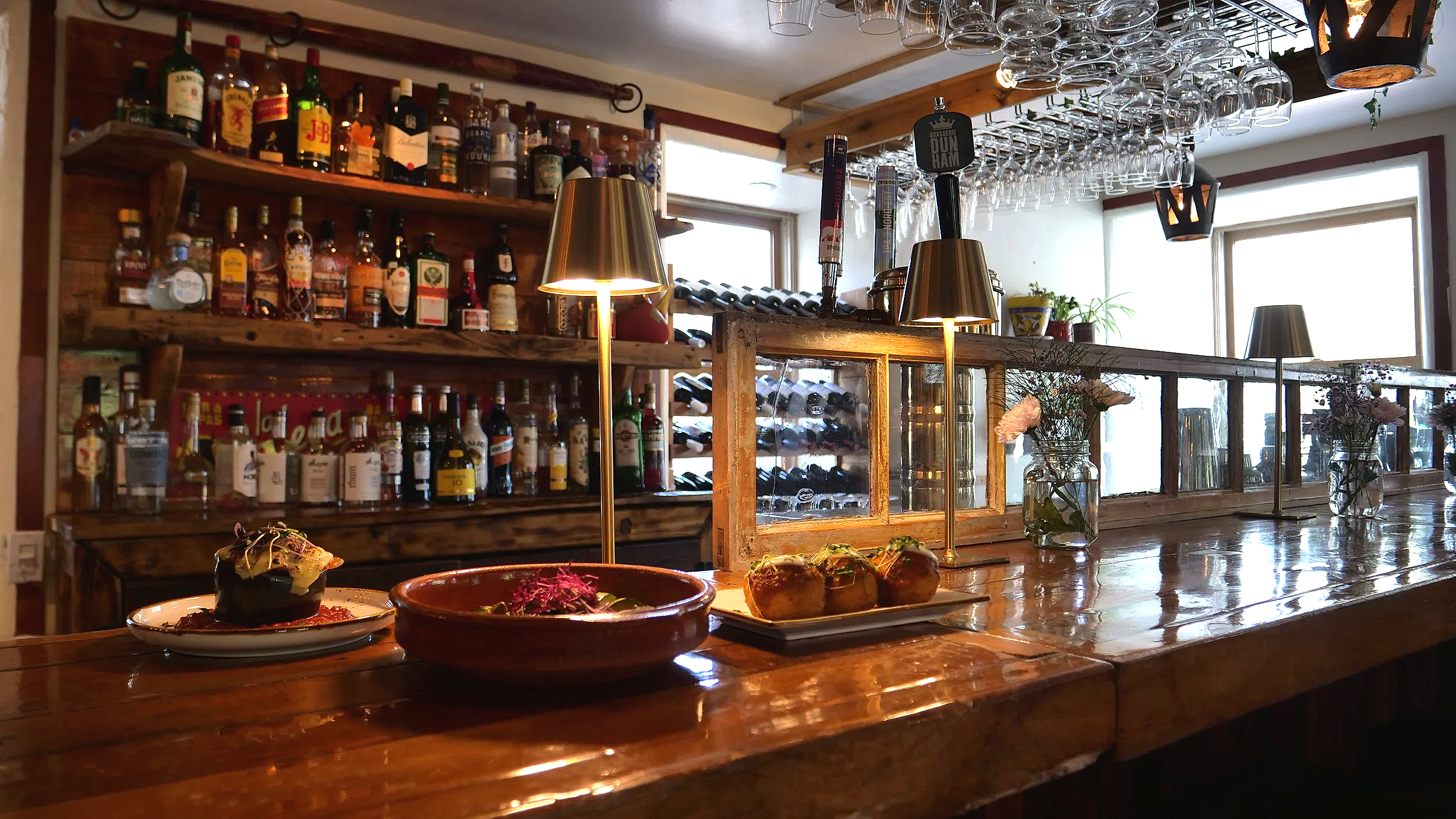 Un bar avec étagères en bois remplies de bouteilles d’alcool, un plafond avec des verres suspendus, et une table en bois avec des plats, fleurs en vases et lampes modernes en métal.