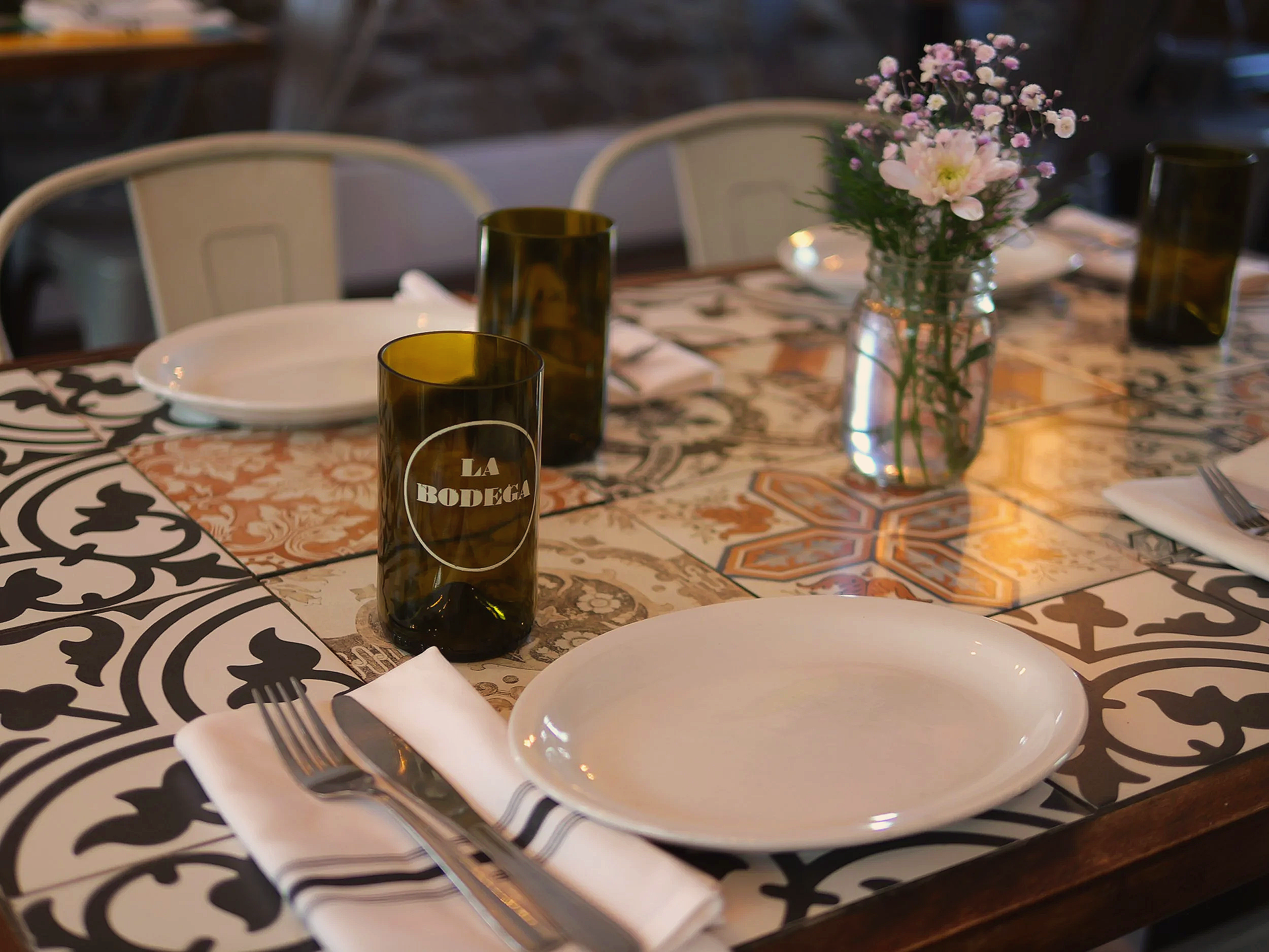 Une table décorée avec des verres en verre teinté marron, des assiettes blanches, des couverts en métal sur des serviettes pliées, un vase avec des fleurs roses, et un napperon à motifs noirs et blancs.