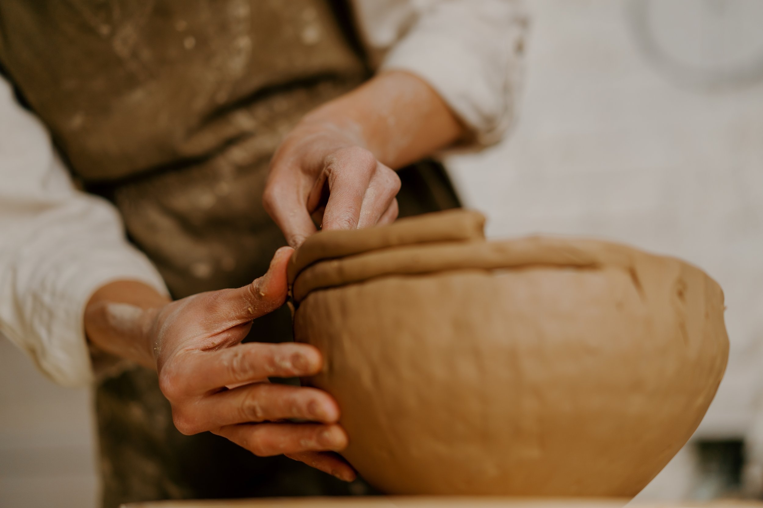 Close-up of a person's hands shaping clay on a pottery wheel, with their body and part of the pottery wheel visible in the background.
