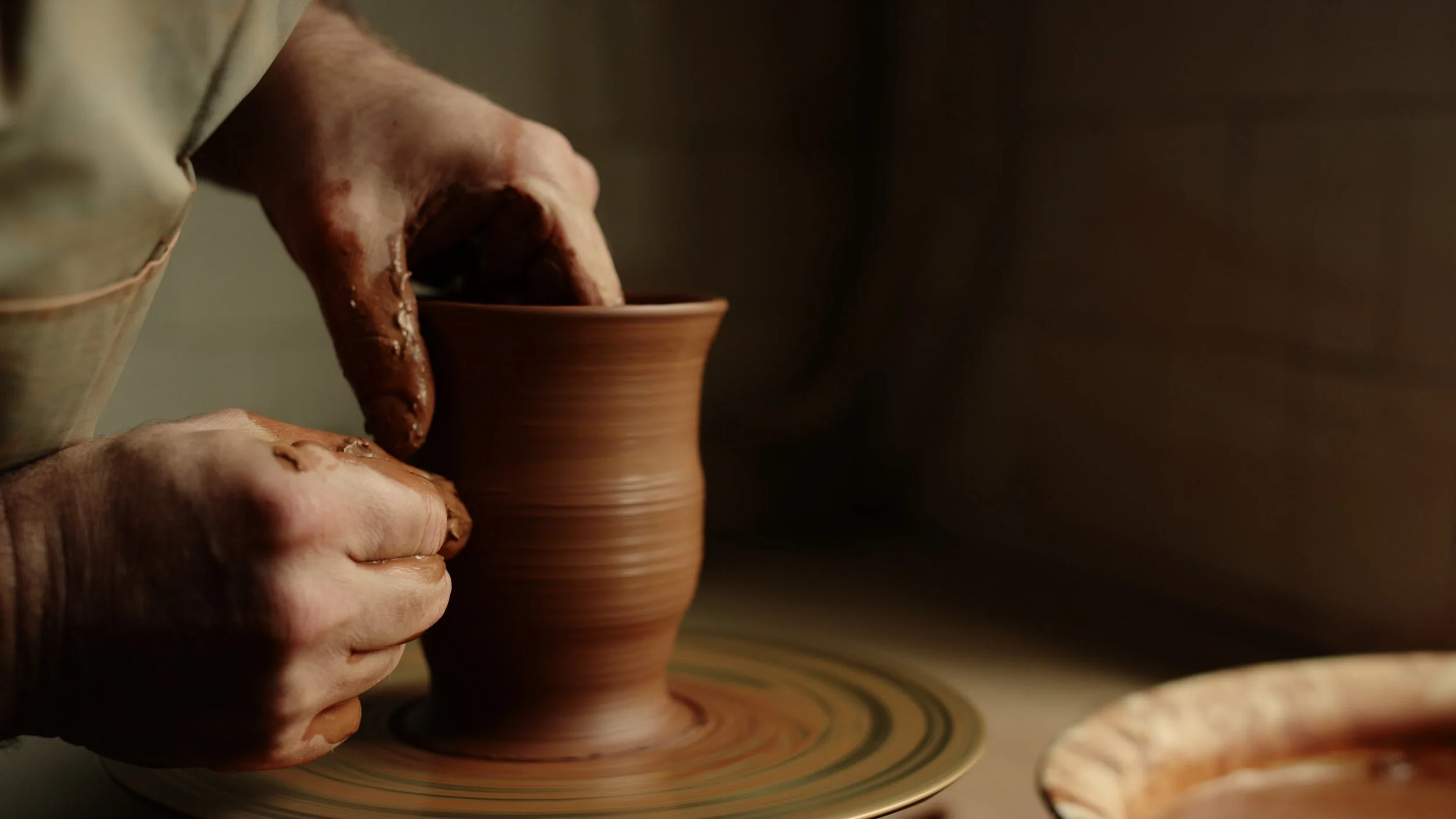 Hands shaping clay on a pottery wheel, with a partially formed clay pot in progress.
