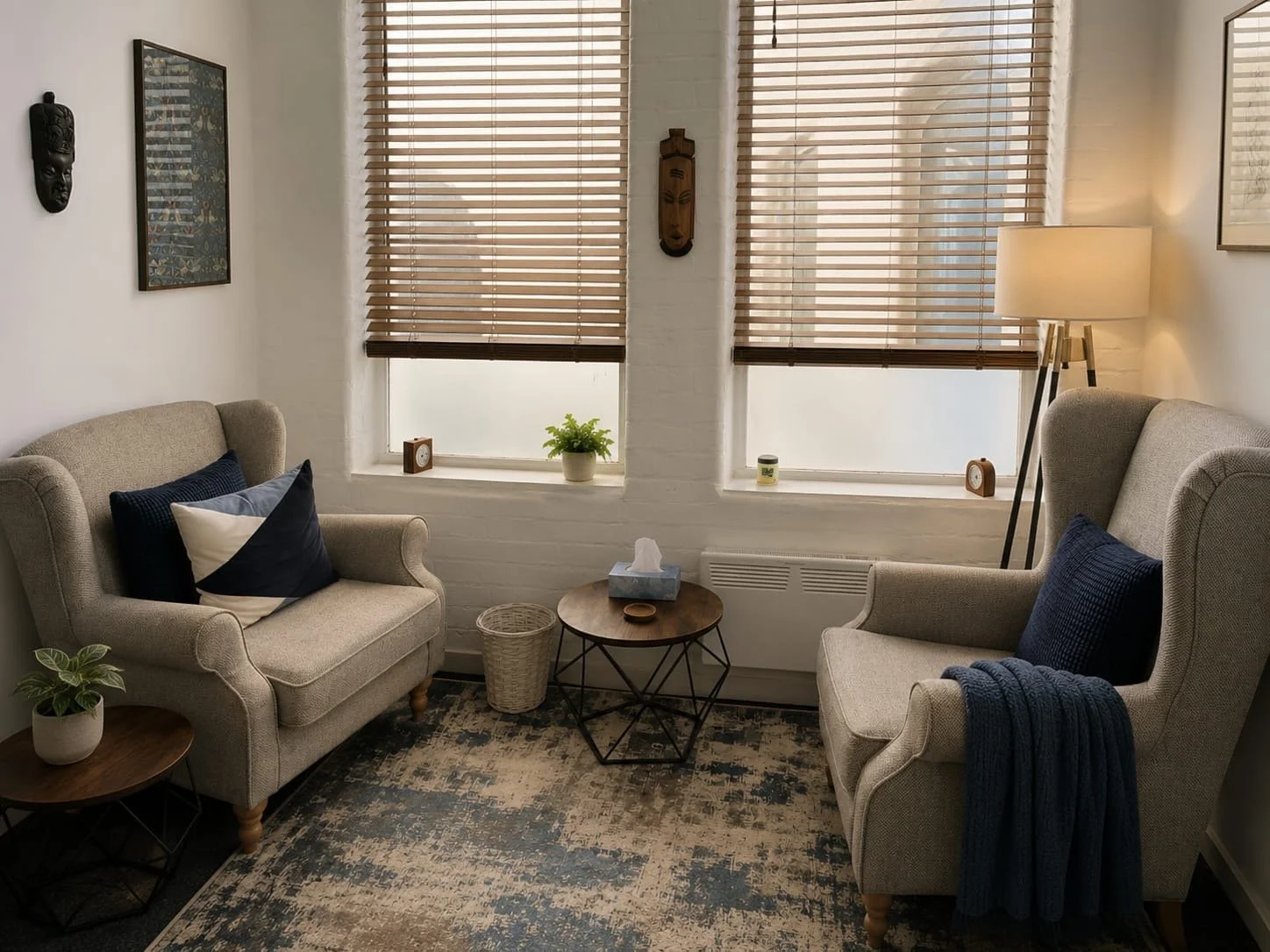 A cozy living room with two beige armchairs, a small round wooden coffee table, and a side table with a potted plant. Windows with wooden blinds let in natural light, and a lamp provides additional lighting. Wall decor includes masks and artwork.