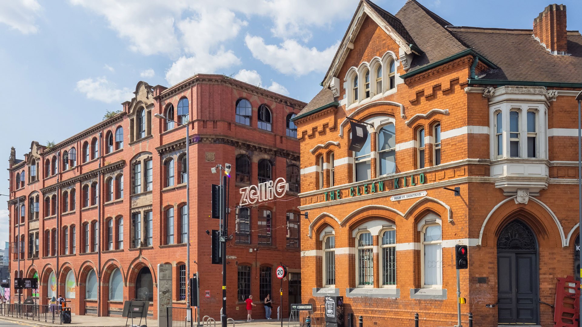 Street view of historic red brick buildings with large arched windows, traffic lights, and pedestrians on sidewalk against partly cloudy sky.