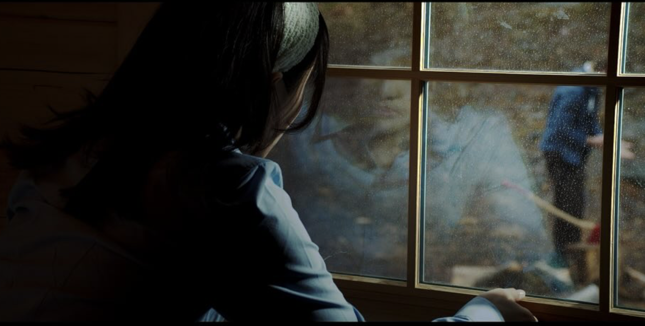 A woman looking out a rain-covered window, with a reflection of her face visible on the glass, while a man in a blue shirt planting a tree outside.