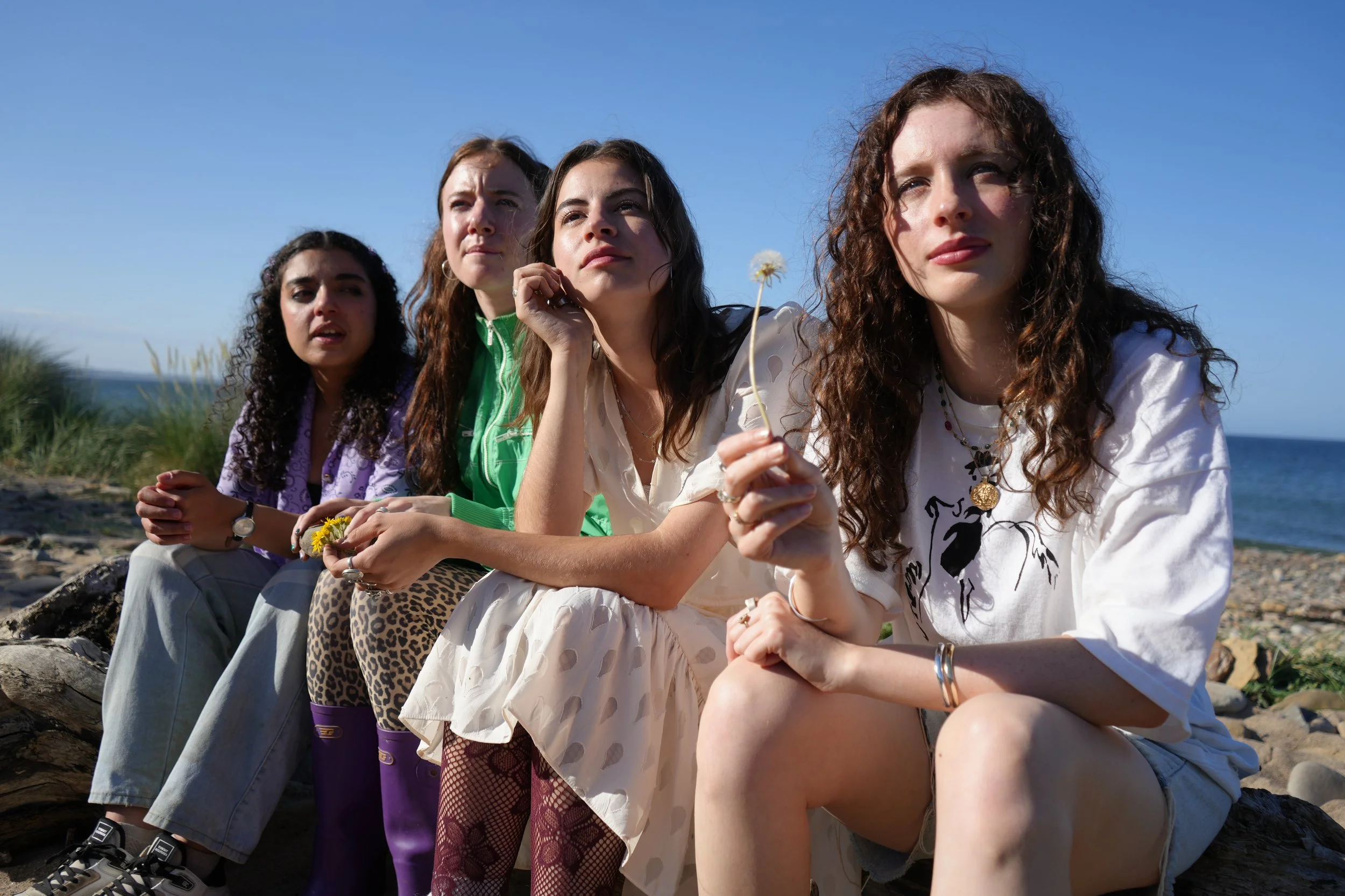 Four young women sitting outdoors on rocks near a beach, looking thoughtfully into the distance against a clear blue sky.