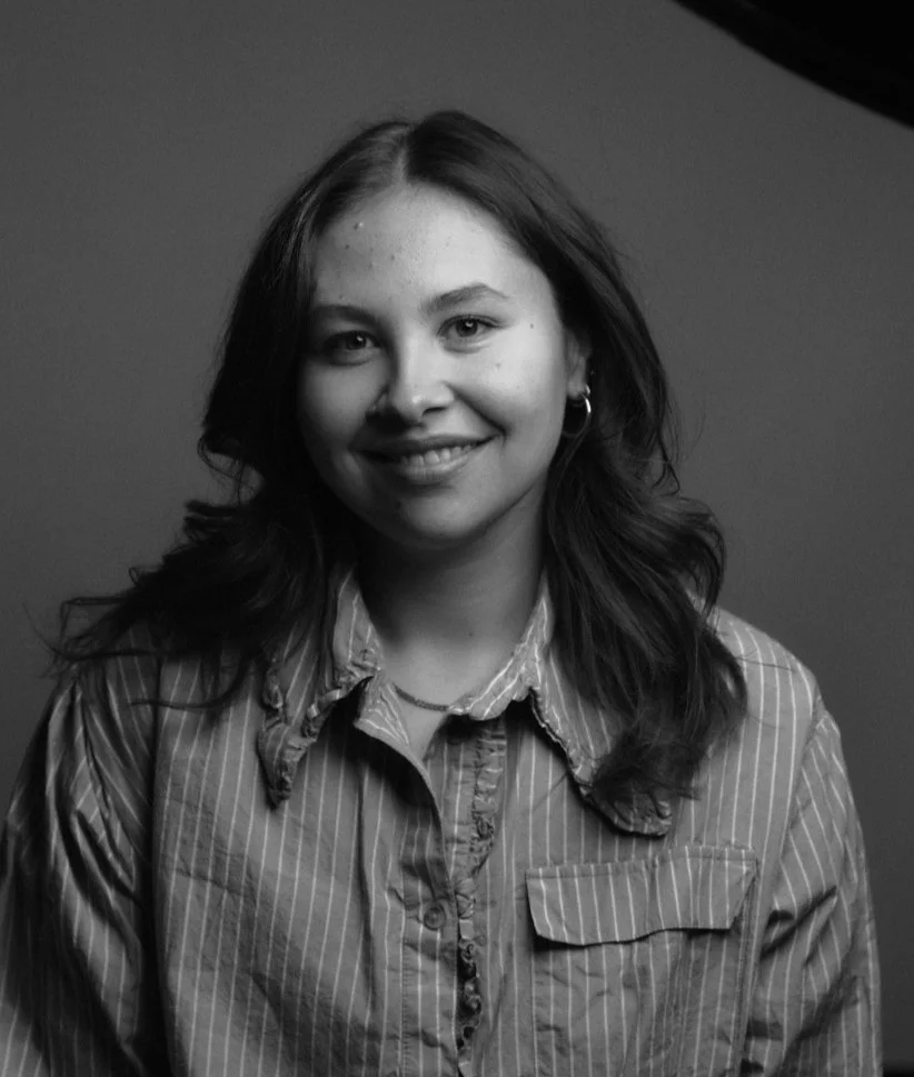 A young woman with shoulder-length wavy hair smiling at the camera, wearing a striped button-up shirt and hoop earrings.