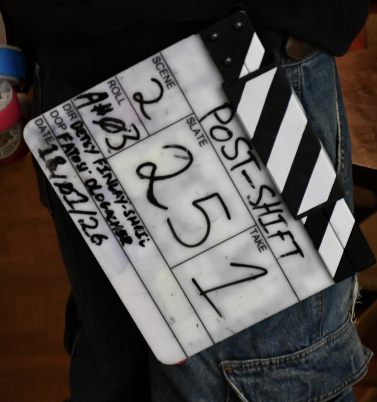 A film slate with the word "POST-SHIFT" written on it, someone wearing jeans sitting nearby, and a partially visible roll of pink tape on the side.
