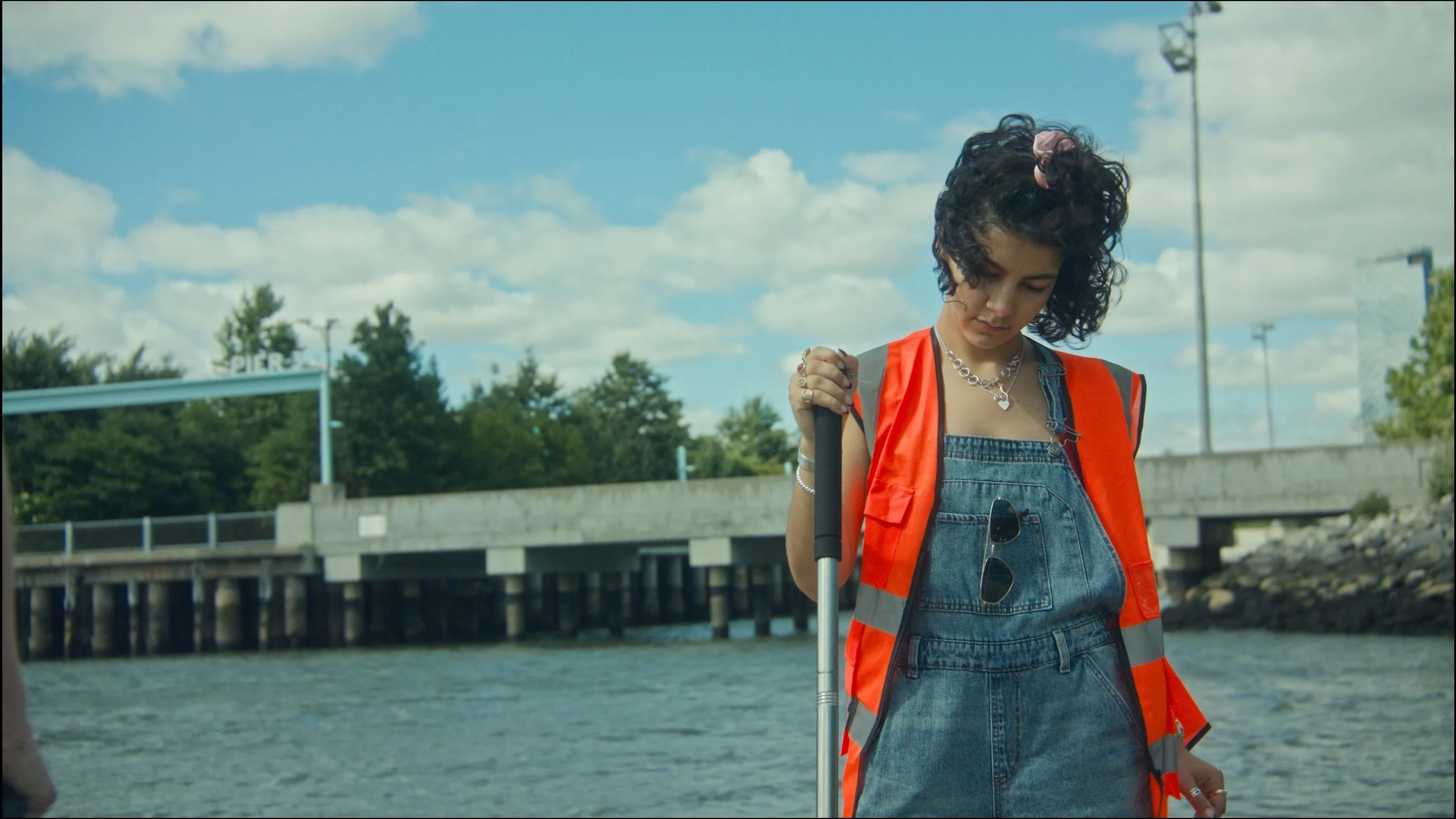 Young woman with curly hair wearing a bright orange safety vest and denim outfit, holding an umbrella, standing outdoors near a waterway with trees and bridge in background under a cloudy sky.