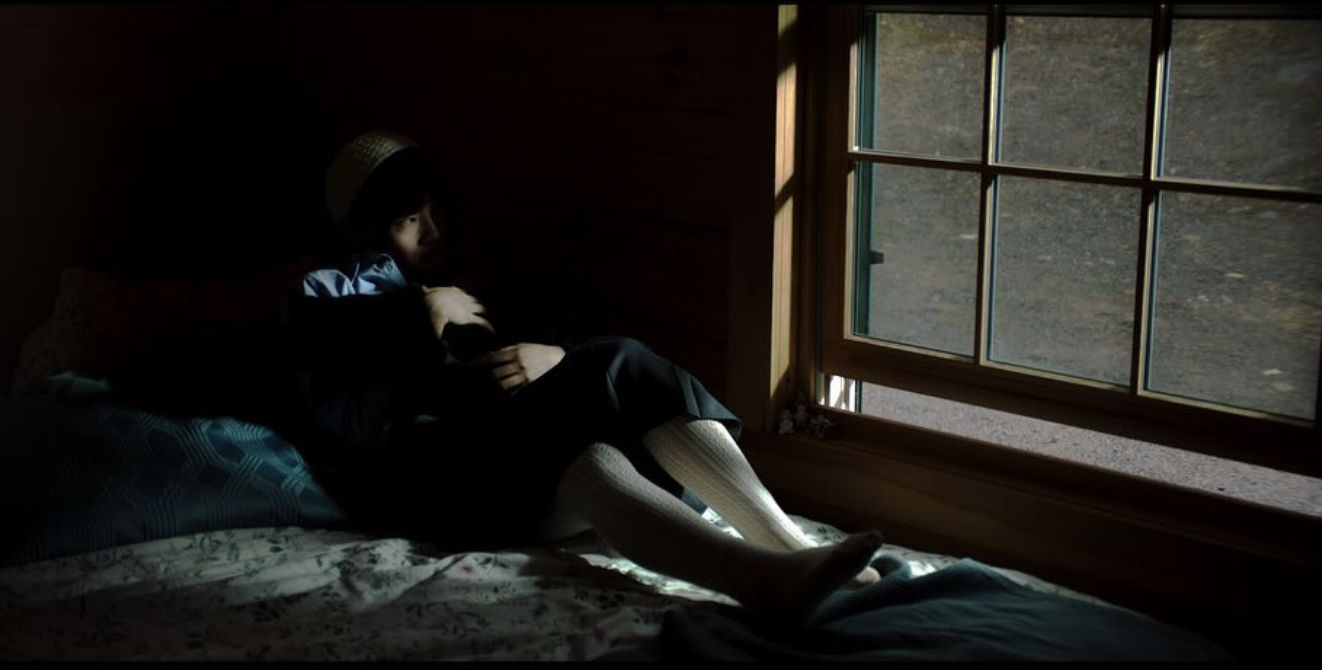 A young girl sitting on a bed near a window, wearing a hat, looking outside to a barren landscape.