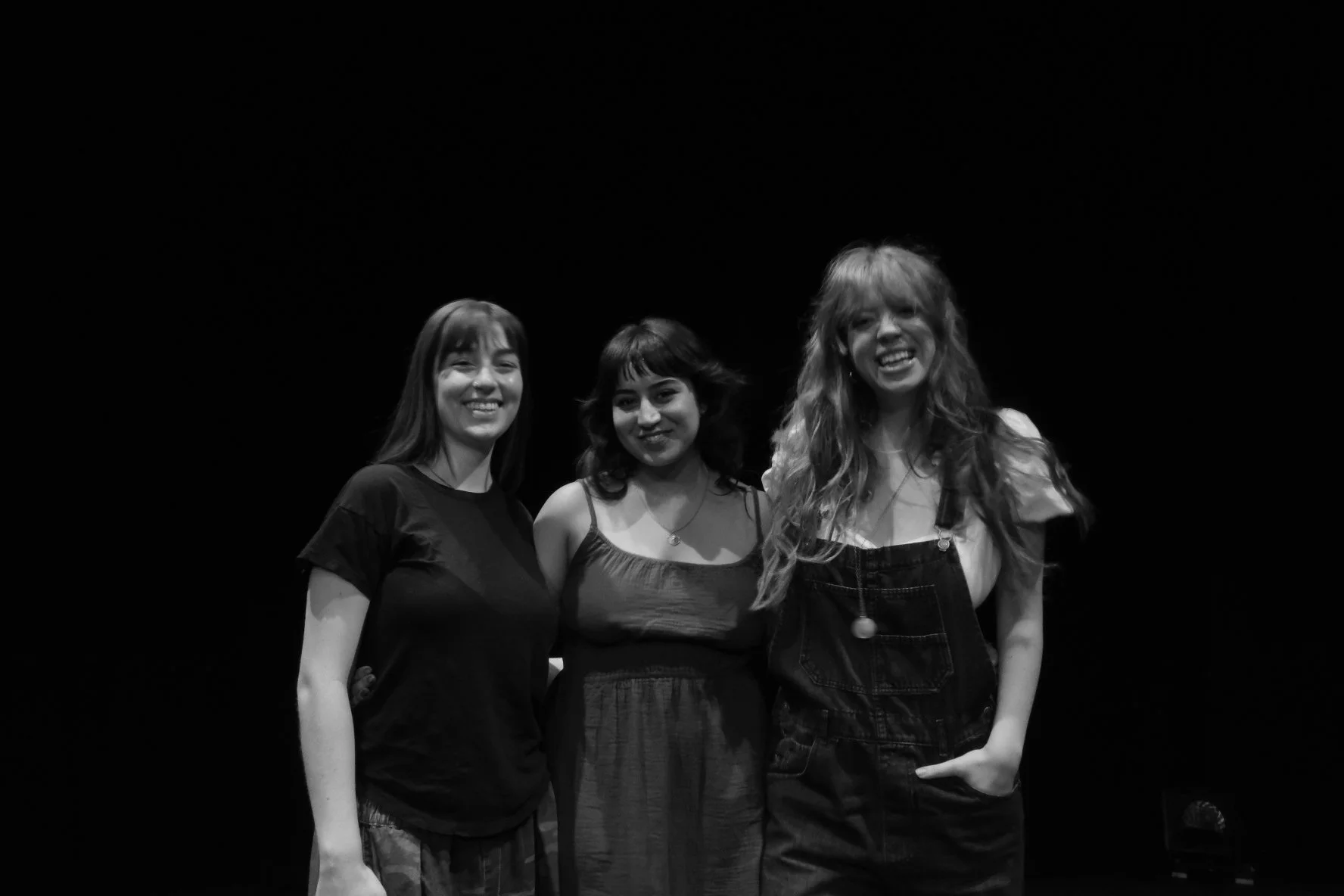Three women standing together smiling against a black background, with the woman on the right wearing overalls with a shirt underneath.