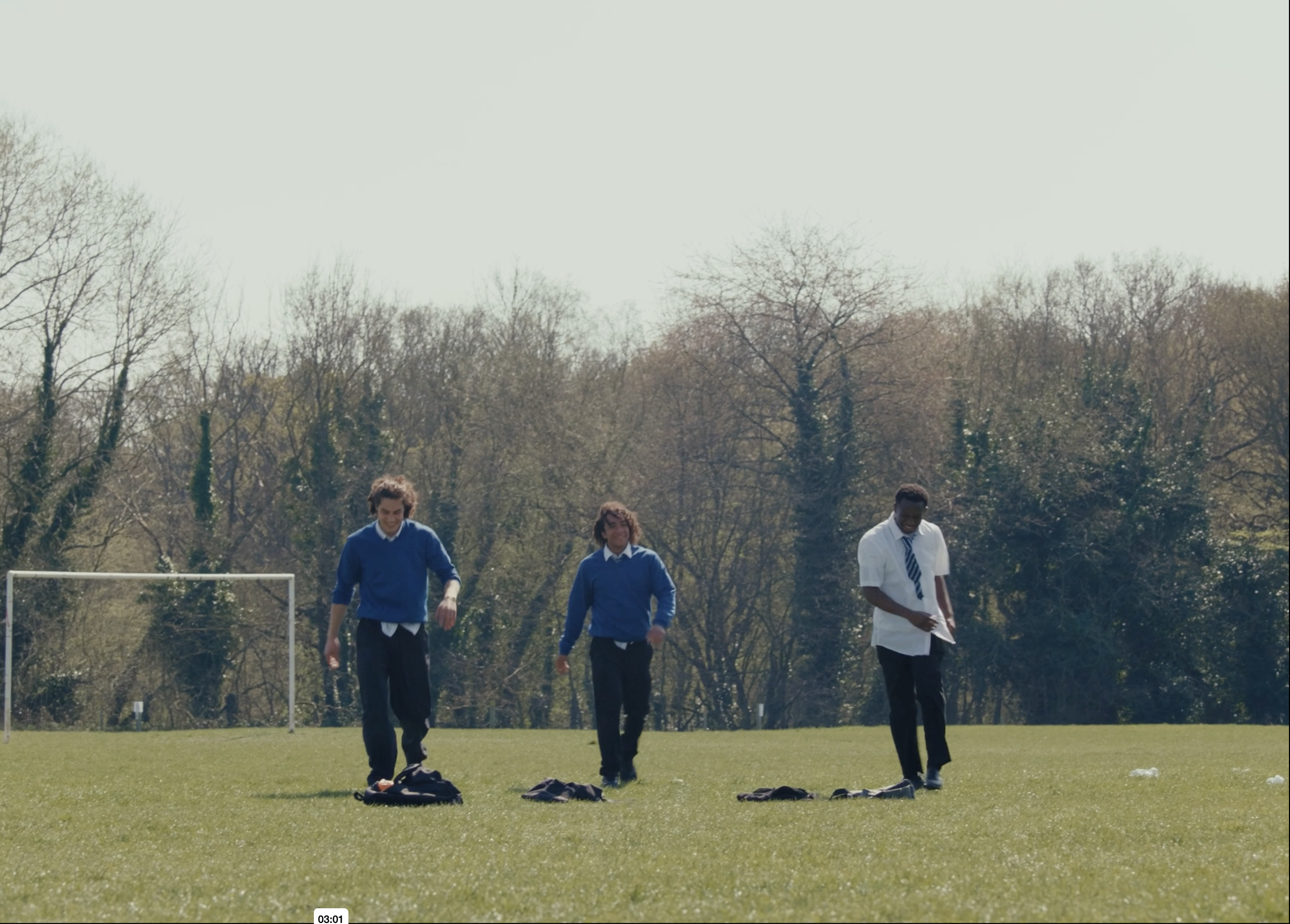 Three young men are walking on a soccer field, with a goalpost in the background and trees behind them. They are dressed in casual and school uniform attire, and there are backpacks on the ground in front of them.