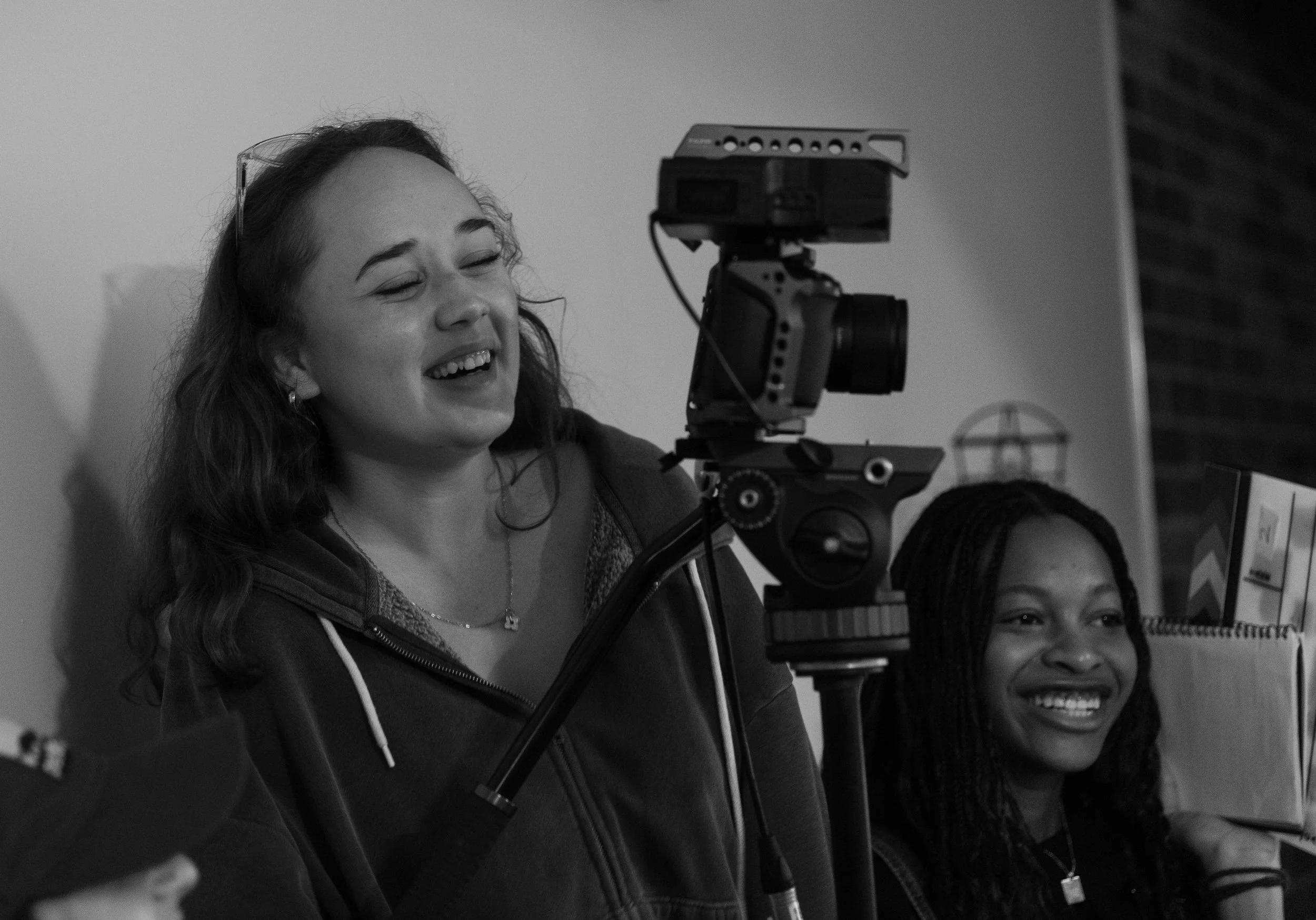 Two women smiling as they look at a camera setup on a tripod.