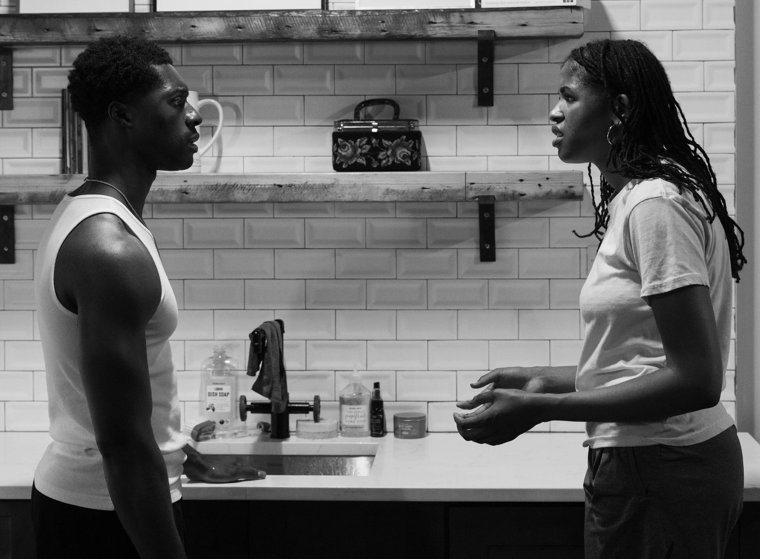 Two women stand facing each other in a kitchen, engaged in a conversation or interaction. The woman on the left has short, curly hair and is wearing a sleeveless top, while the woman on the right has long, dreadlocked hair and is wearing a T-shirt. A shelf with various bottles and a decorative case are visible in the background.