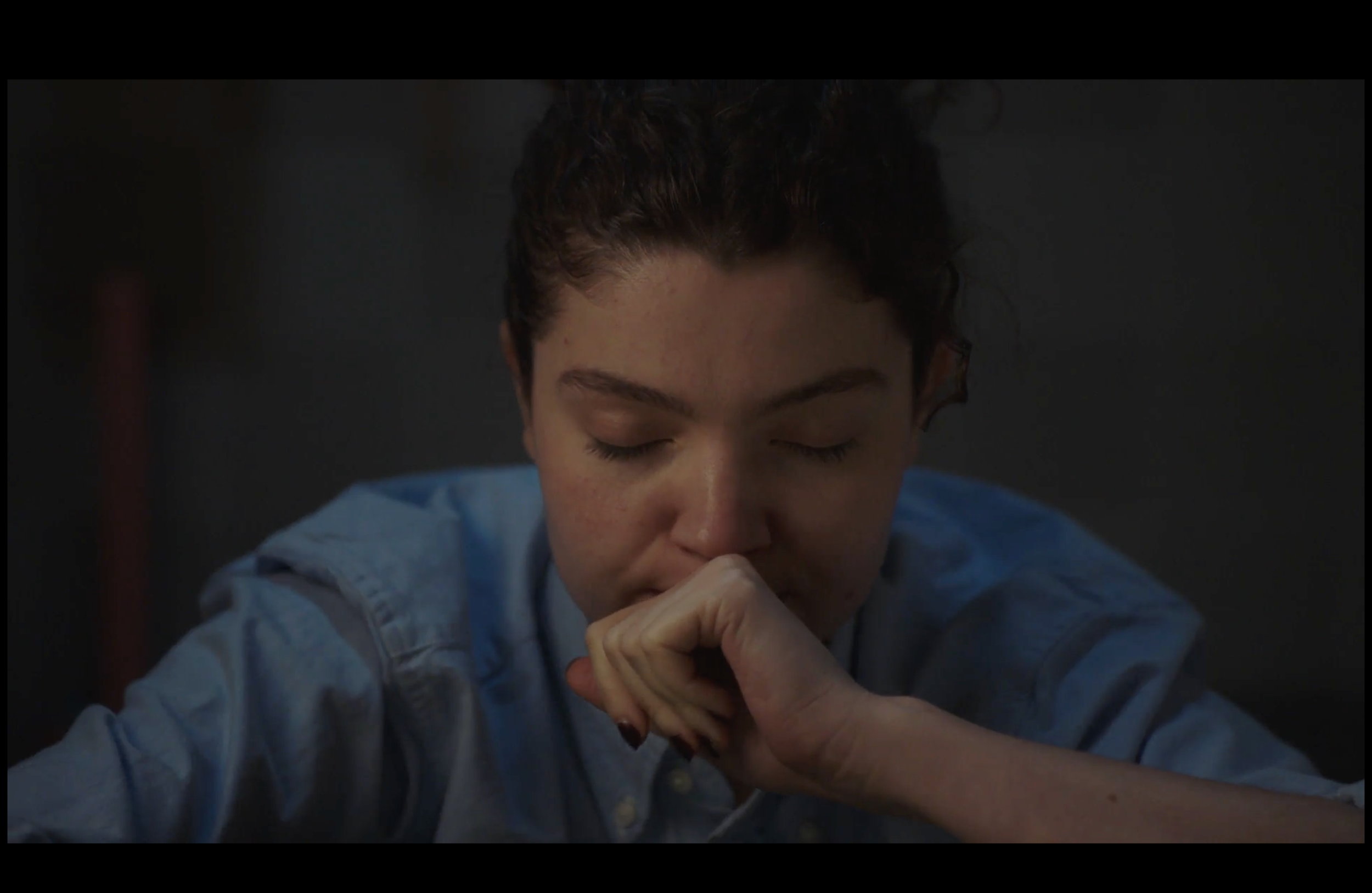A young person with curly hair and closed eyes, resting their chin on their hand, wearing a blue shirt, in a dark environment.
