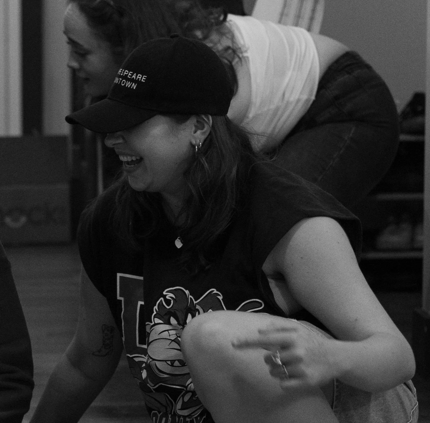A woman with long hair, wearing a baseball cap and a graphic t-shirt, sitting on the floor and smiling.