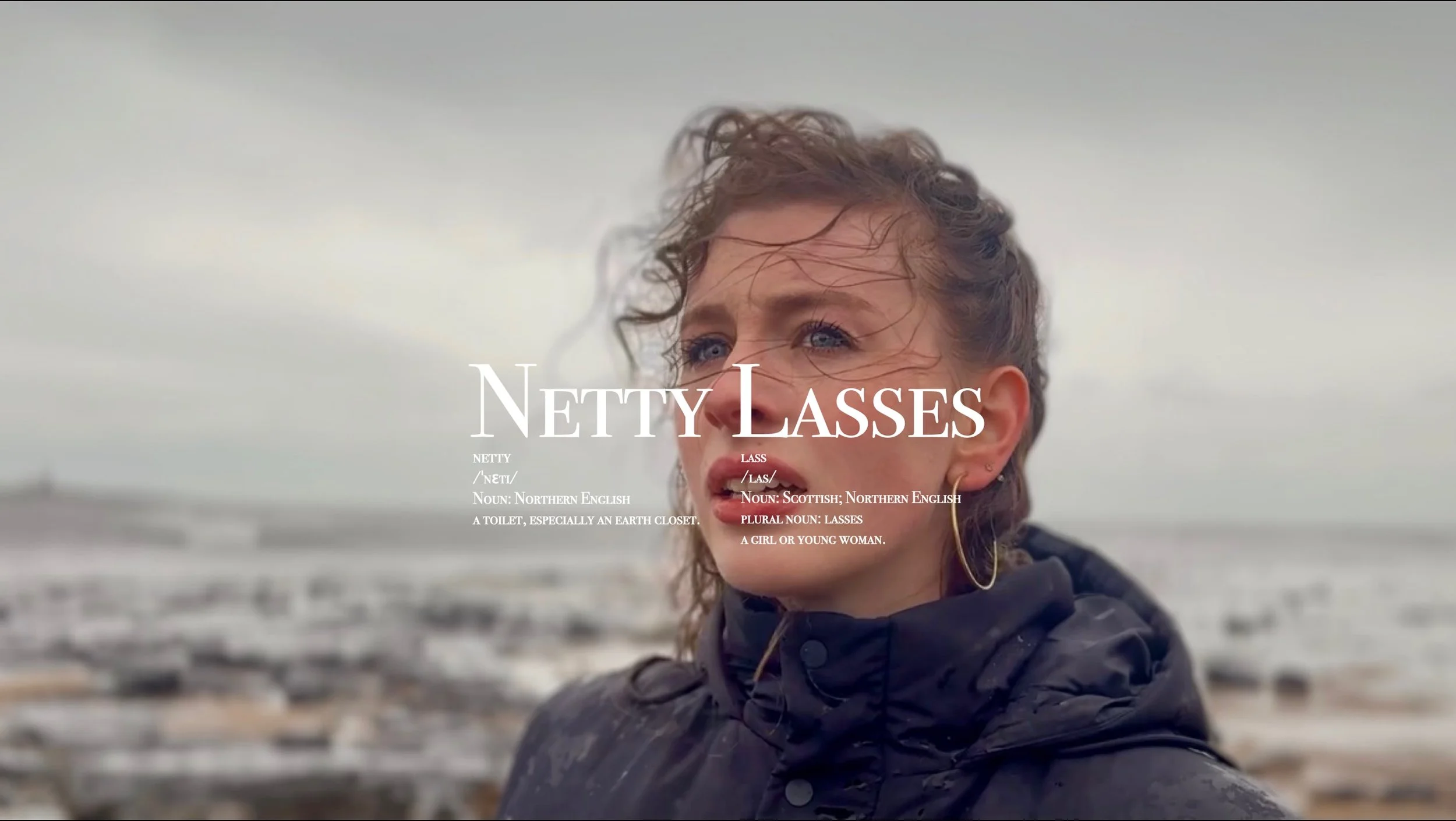 Close-up of a young woman with curly hair and blue eyes, standing outdoors on a cloudy day at the beach, looking into the distance. Overlaid text defines the word 'netty' as a Northern English slang term for a toilet, especially an earth closet, and 'lass' as a Scottish Northern English term for a girl or young woman.