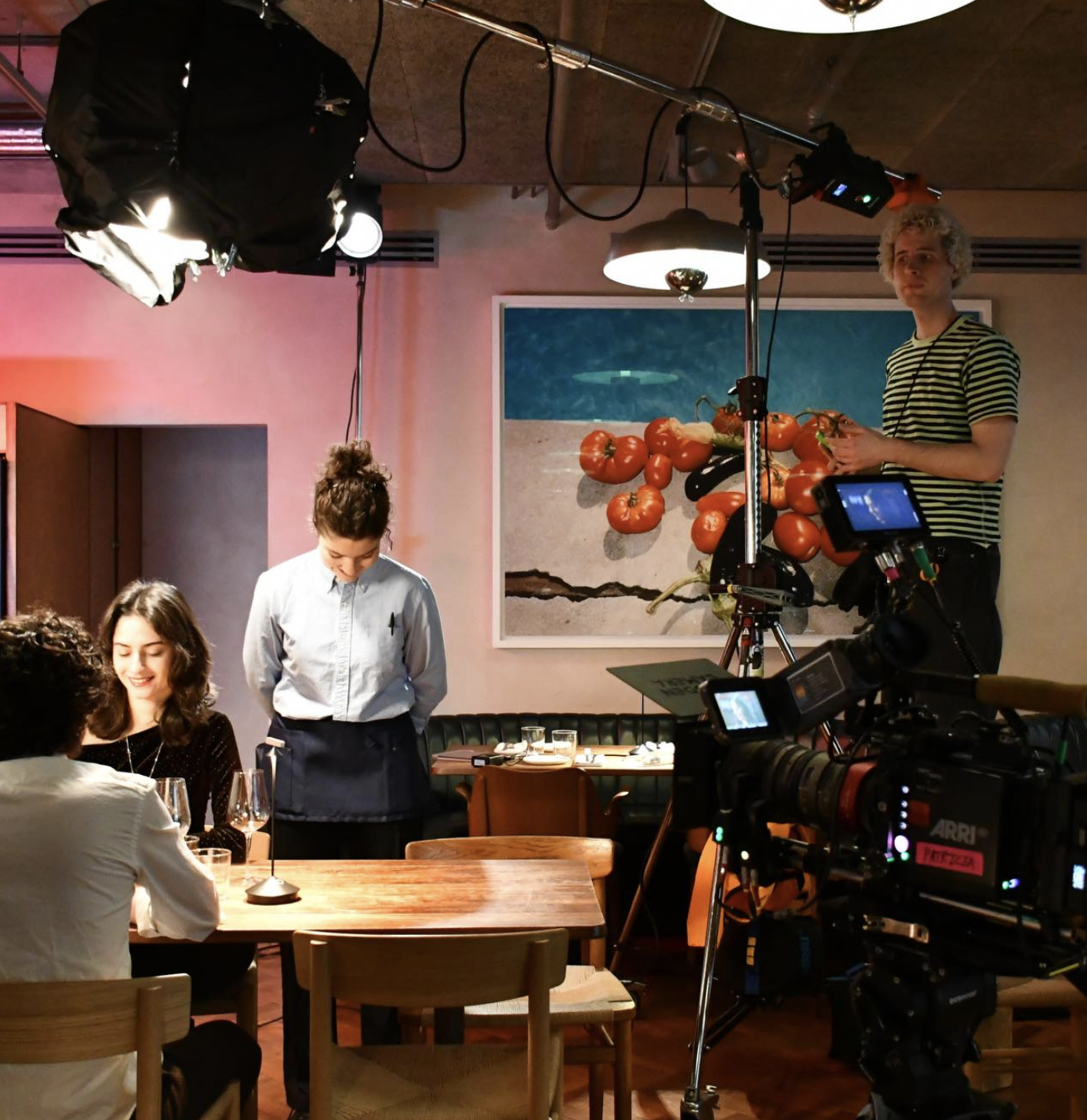 Filming crew setting up a scene in a restaurant with actors seated at a table, a waitress standing by, and professional camera and lighting equipment.