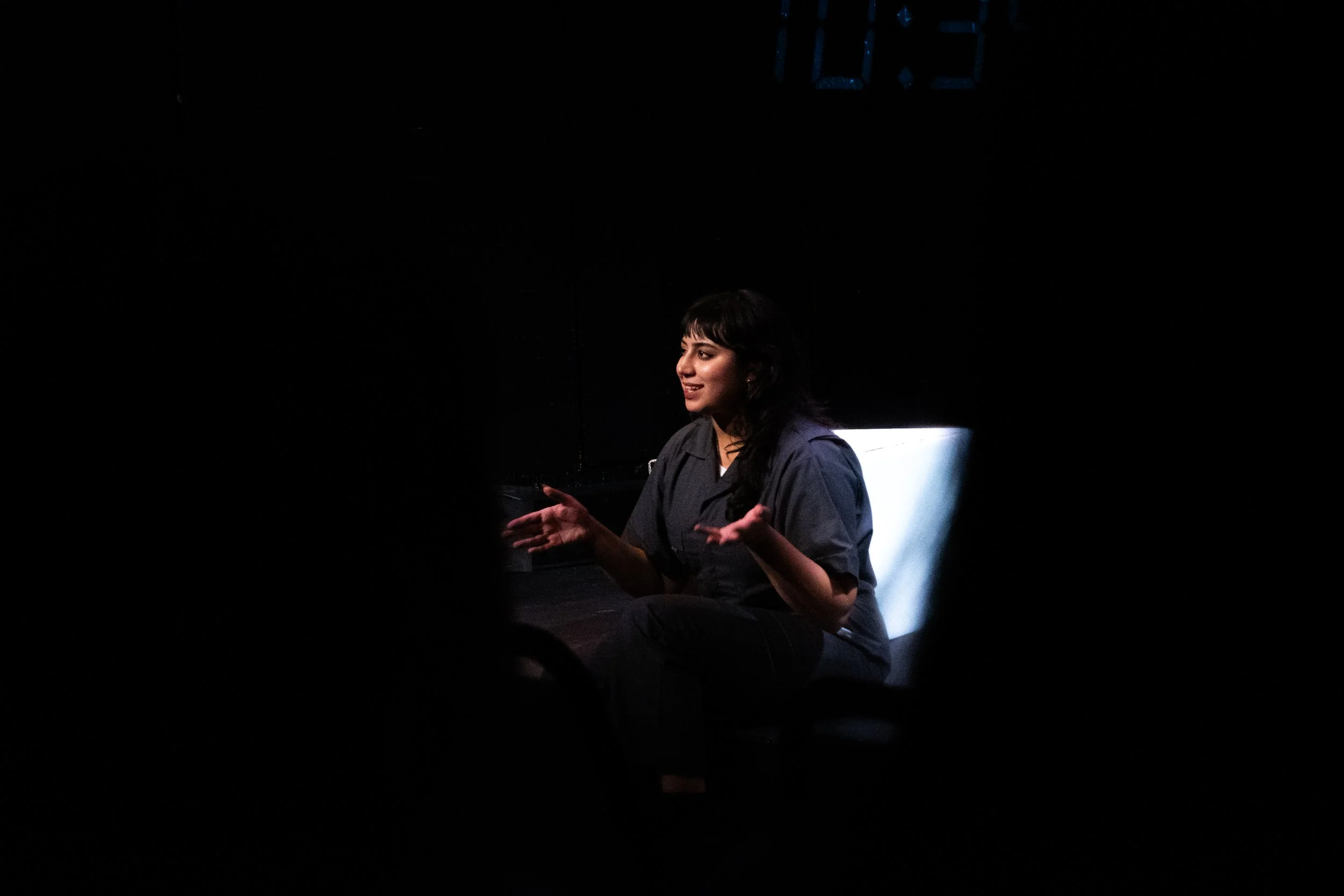 A woman with dark hair, wearing a dark shirt, is speaking or performing on a dimly lit stage. A bright light source is illuminating her from the side. There is a digital clock showing 12:21 in the background.