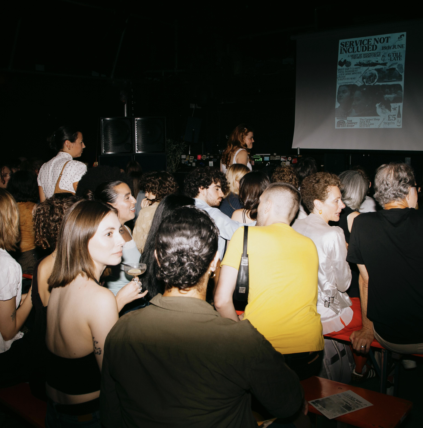 People sitting and standing in a dark theater or concert venue, watching a presentation on a large screen.